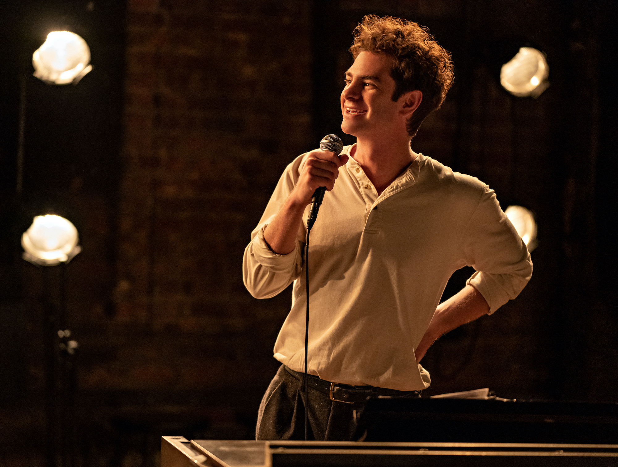 Andrew Garfield as Jonathan Larson in 'TICK, TICK…BOOM!' He stands on a stage with brick walls behind him and warm amber stage lights on the sides. He wears a white long-sleeved shirt with three buttons and dark pants and holds a microphone up to his face as he smiles.