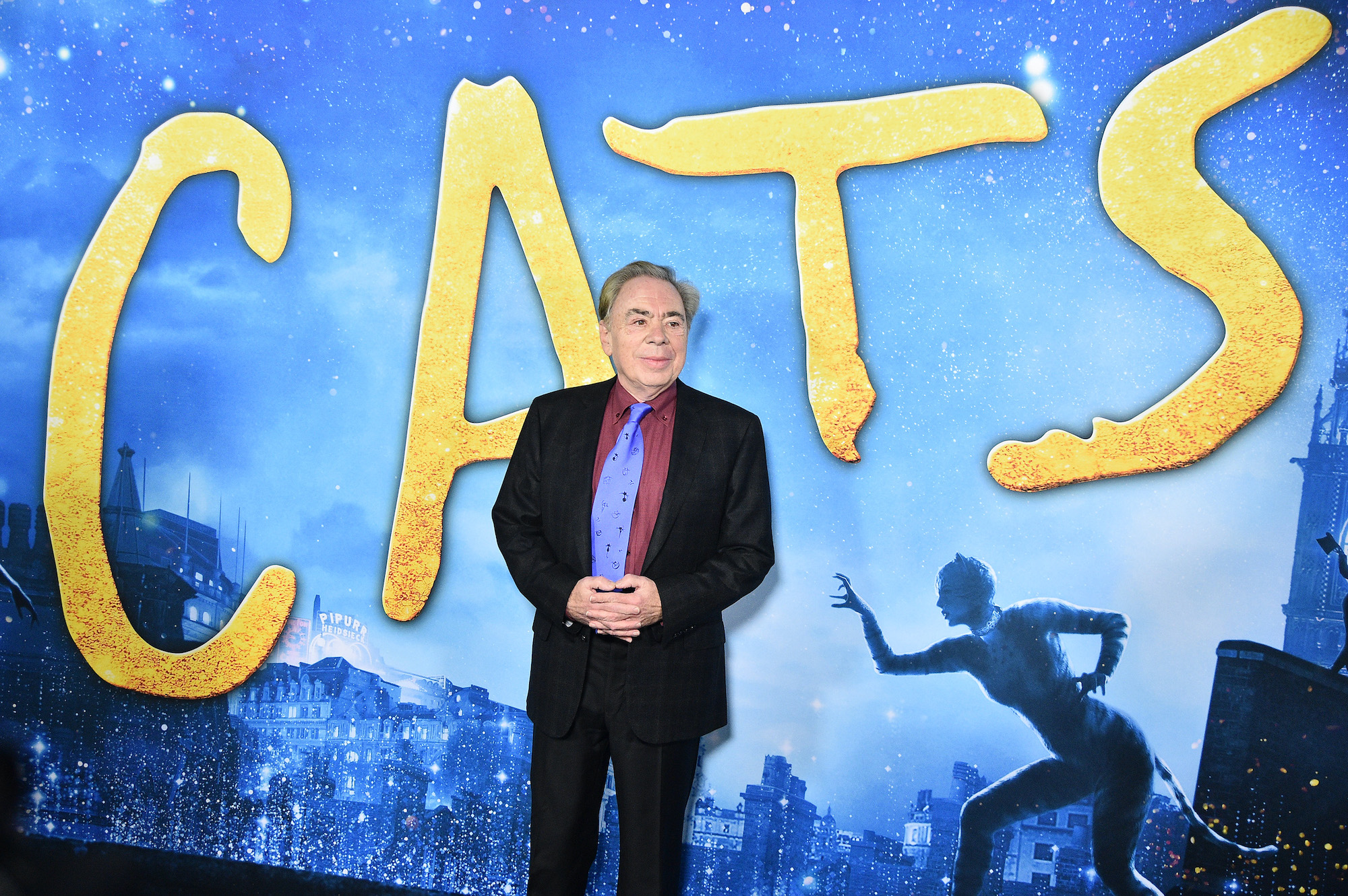 Andrew Lloyd Webber attends the 'Cats' world premiere in New York City in 2019. He poses for photos in a black suit with a red shirt and blue tie and his hands clasped. Behind him is a large blue backdrop that says 'CATS' in big yellow letters.