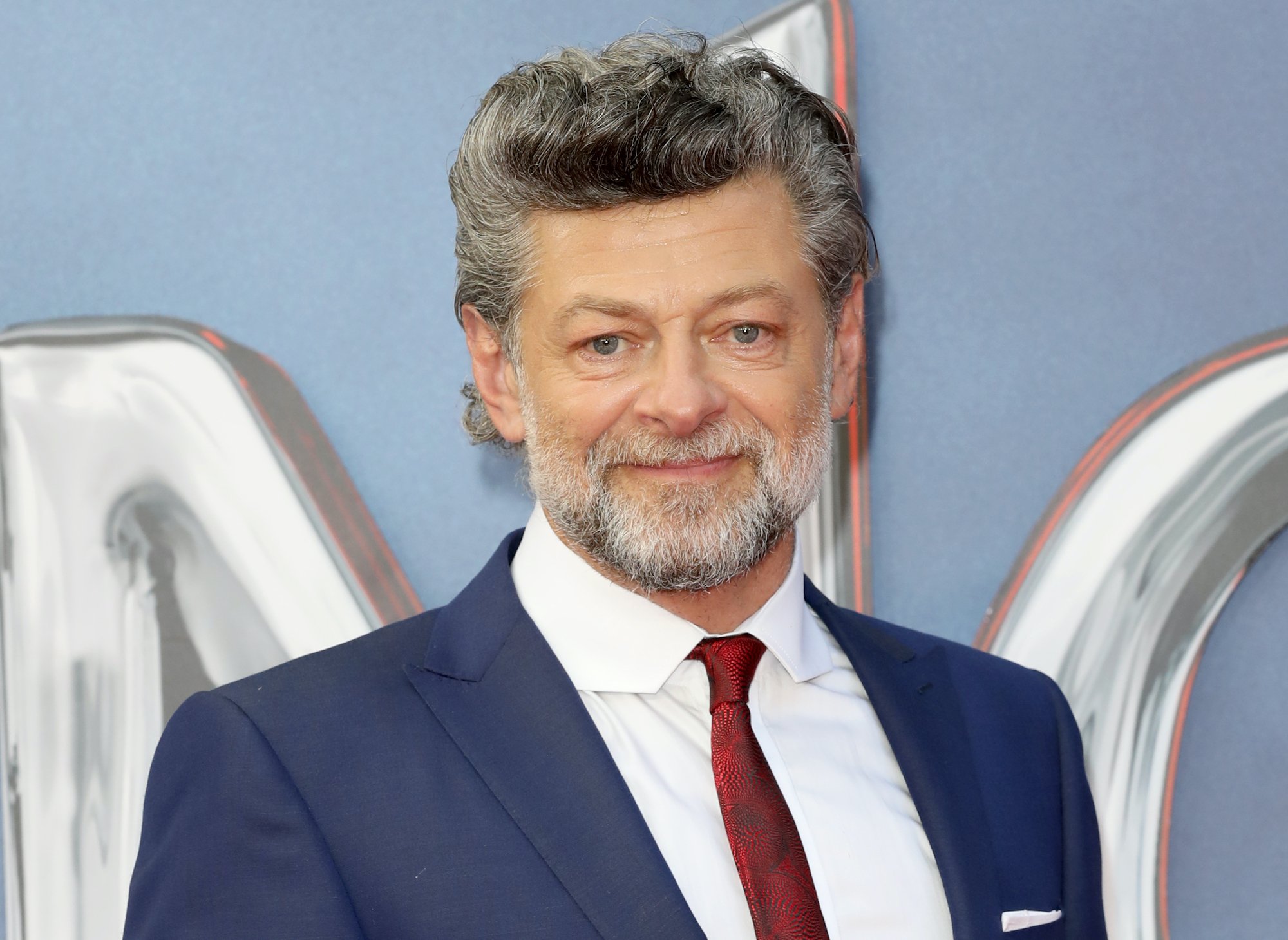 Director Andy Serkis attends the fan screening of 'Venom: Let There Be Carnage' at Cineworld Leicester Square on September 14, 2021 in London, England. He wears a dark blue suit, white shirt, and red tie as he stands in front of the light blue-grey backdrop.