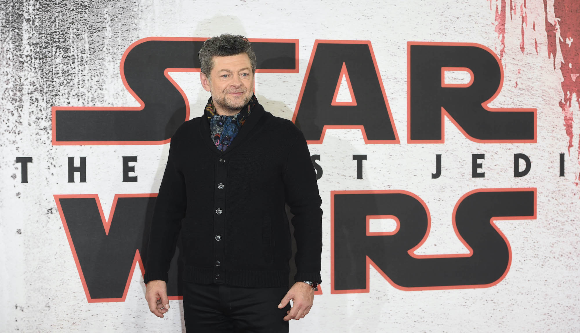 Andy Serkis during the 'Star Wars: The Last Jedi' photo call on Dec. 13, 2017 in London, England. He stands in black pants and a black sweater with a blue and purple scarf in front of a white and red backdrop that says 'Star Wars: The Last Jedi.'