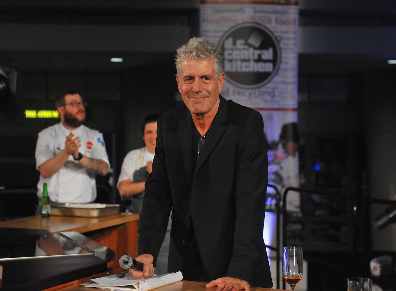 Anthony Bourdain smirks as he wears a black suit, holds a microphone, and looks on