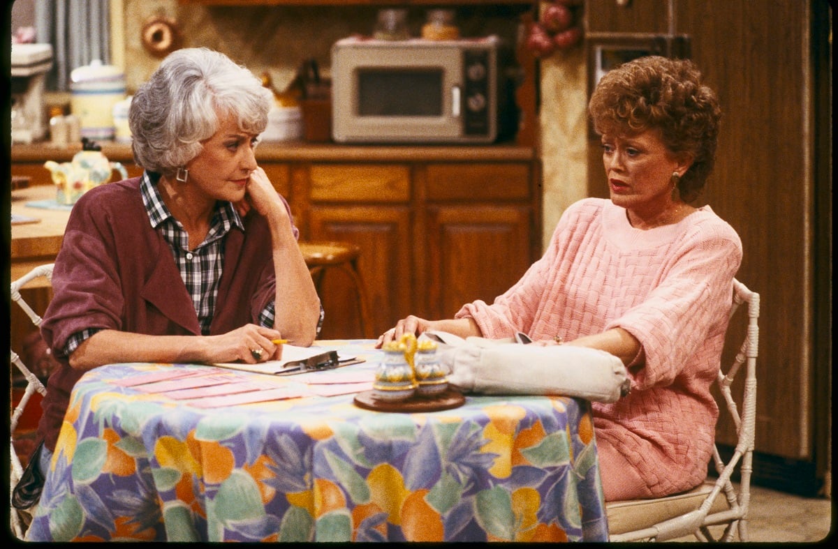'The Golden Girls' actor Bea Arthur in a purple sweater and plaid shirt, and Rue McClanahan in a pink outfit, sitting at the kitchen table.