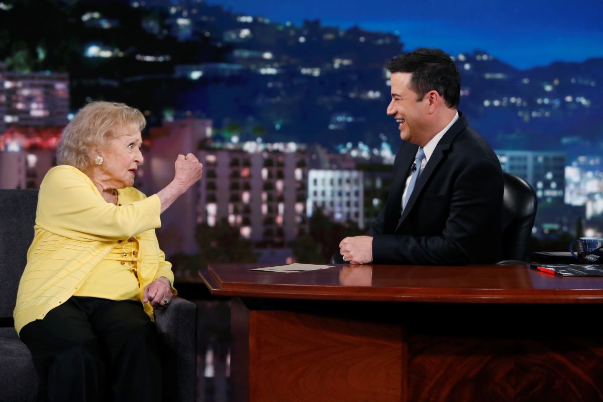 Betty White in yellow and showing a fist to Jimmy Kimmel, smiling and seated behind his desk
