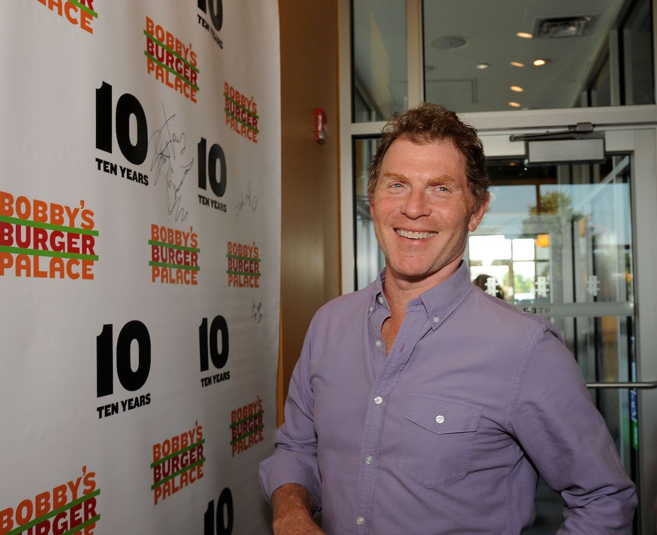 Bobby Flay smiles next to a wall that says 'Bobby's Burger Palace' and '10 years'