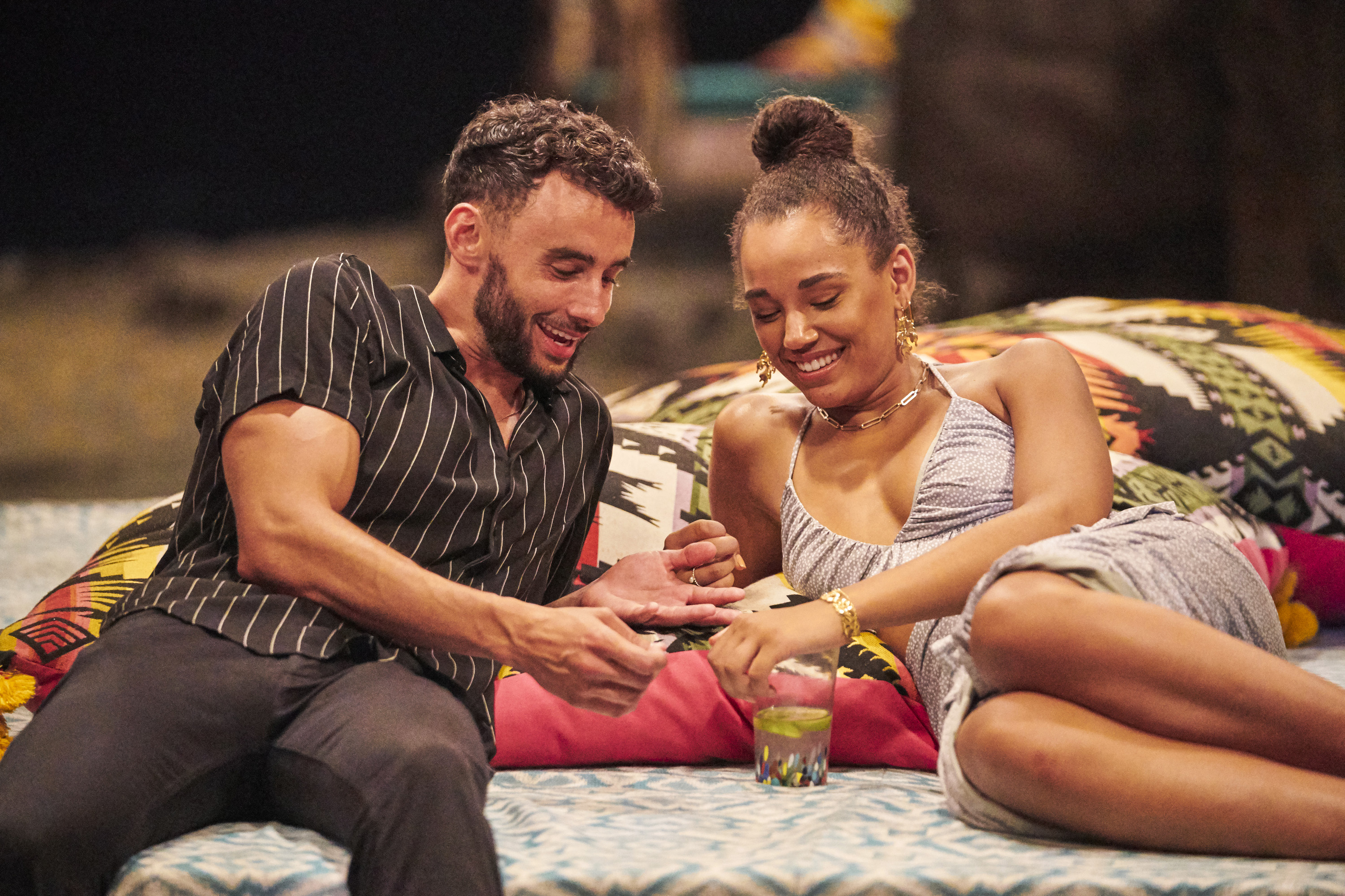 'Bachelor in Paradise' stars Brendan Morais and Pieper James cuddle up on one of the beds on the beach. Brendan wears a black short sleeve button up shirt with thin white stripes and black pants. Pieper wears a gray dress with white polka dots.