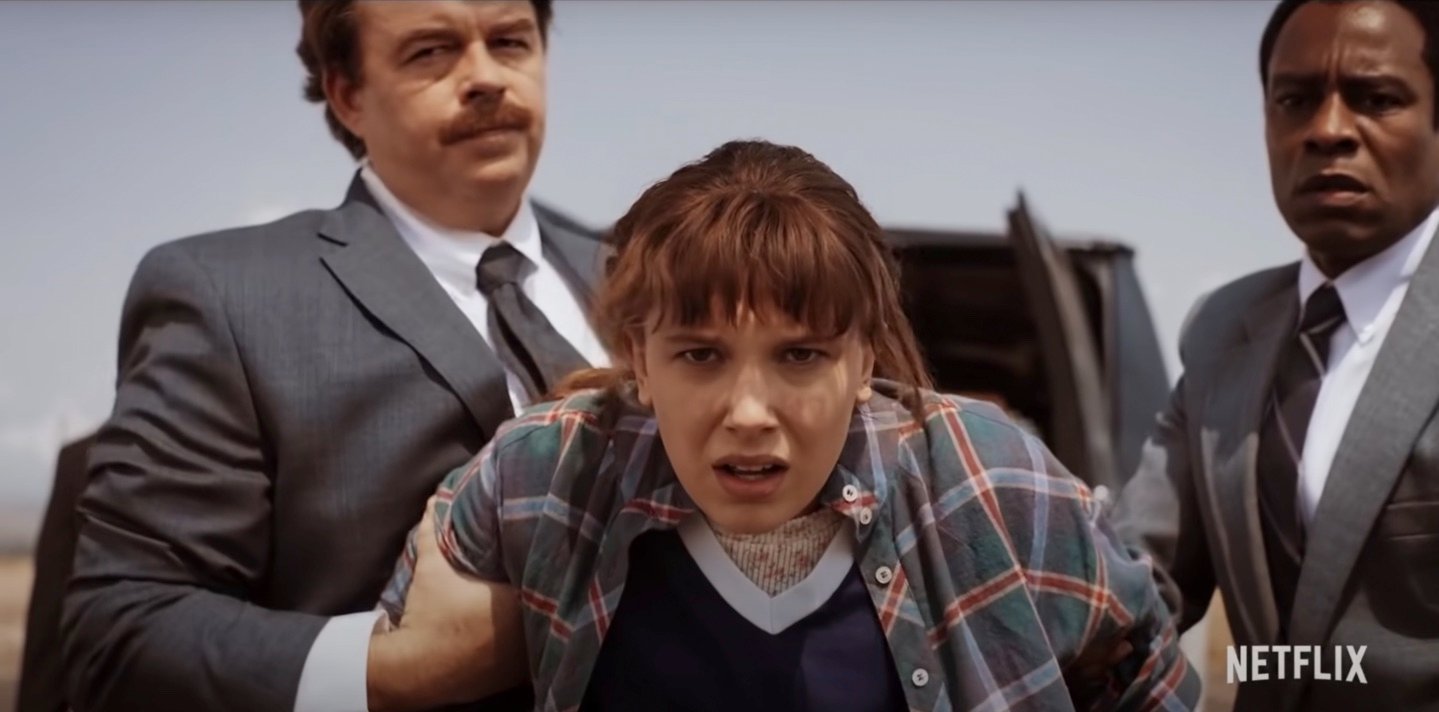 Eleven from 'Stranger Things' Season 4 in a plaid shirt while two men hold her back.