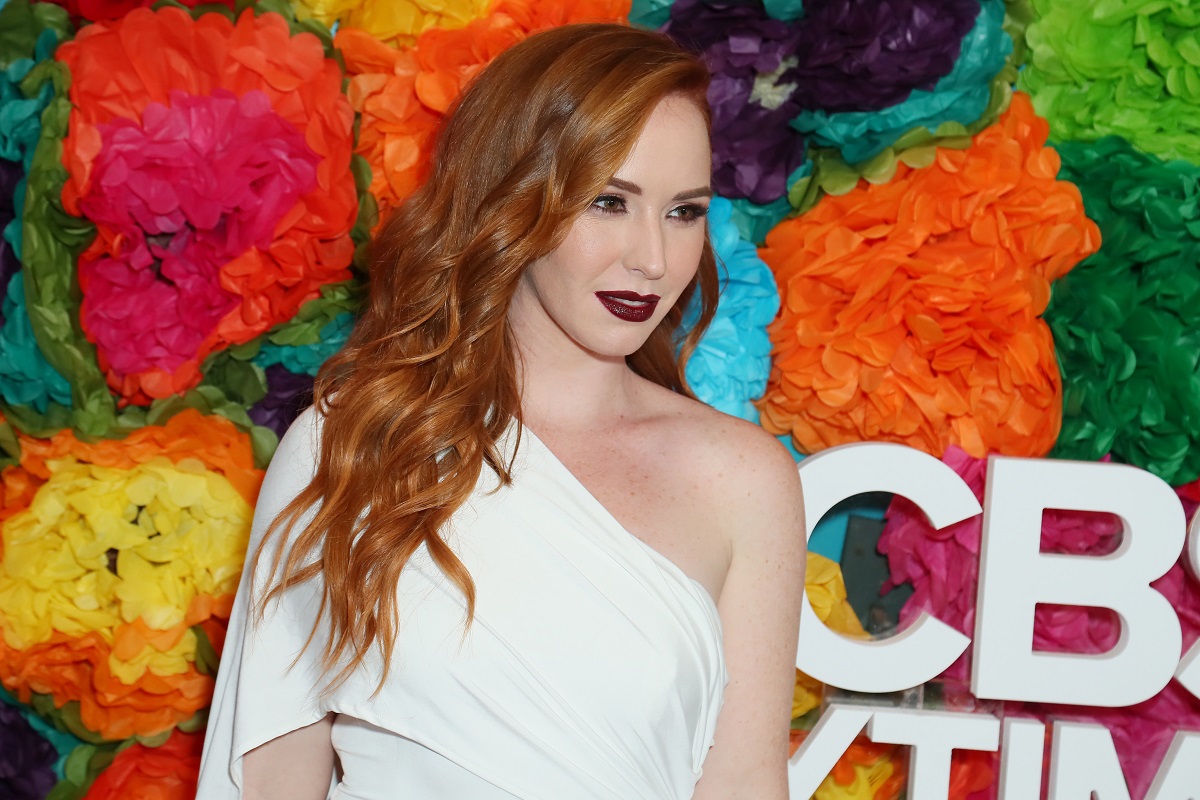 'The Young and the Restless' actor Camryn Grimes wearing a white dress and dark lipstick, and standing in front of a floral background.