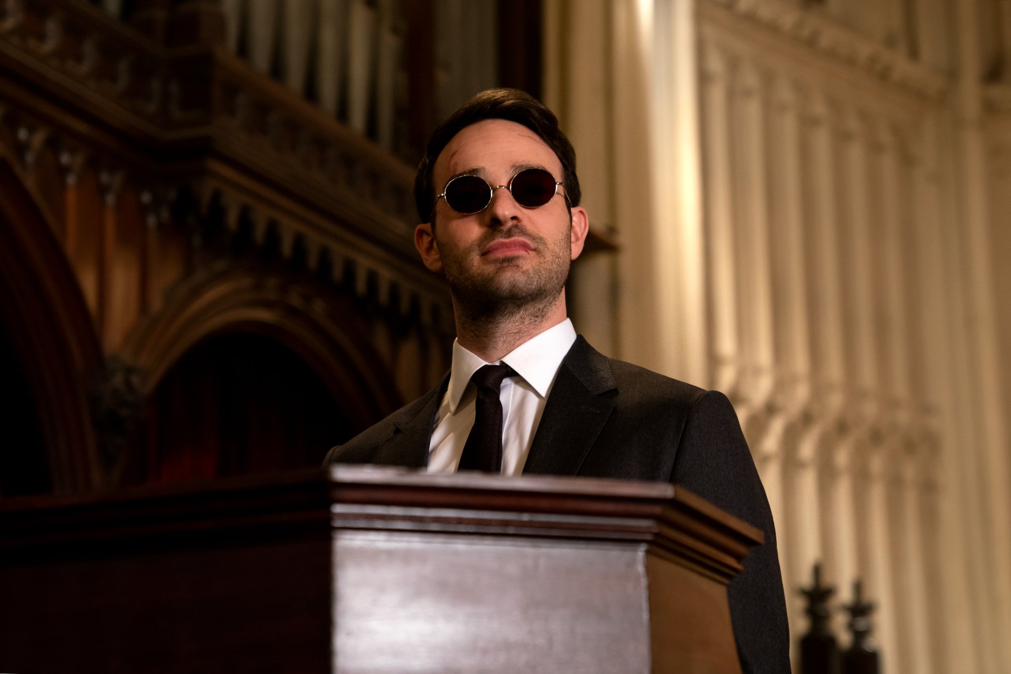 Charlie Cox as Matt Murdock/Daredevil in Netflix's 'Daredevil' series. He's wearing a suit and standing at a podium. He has dark glasses on.