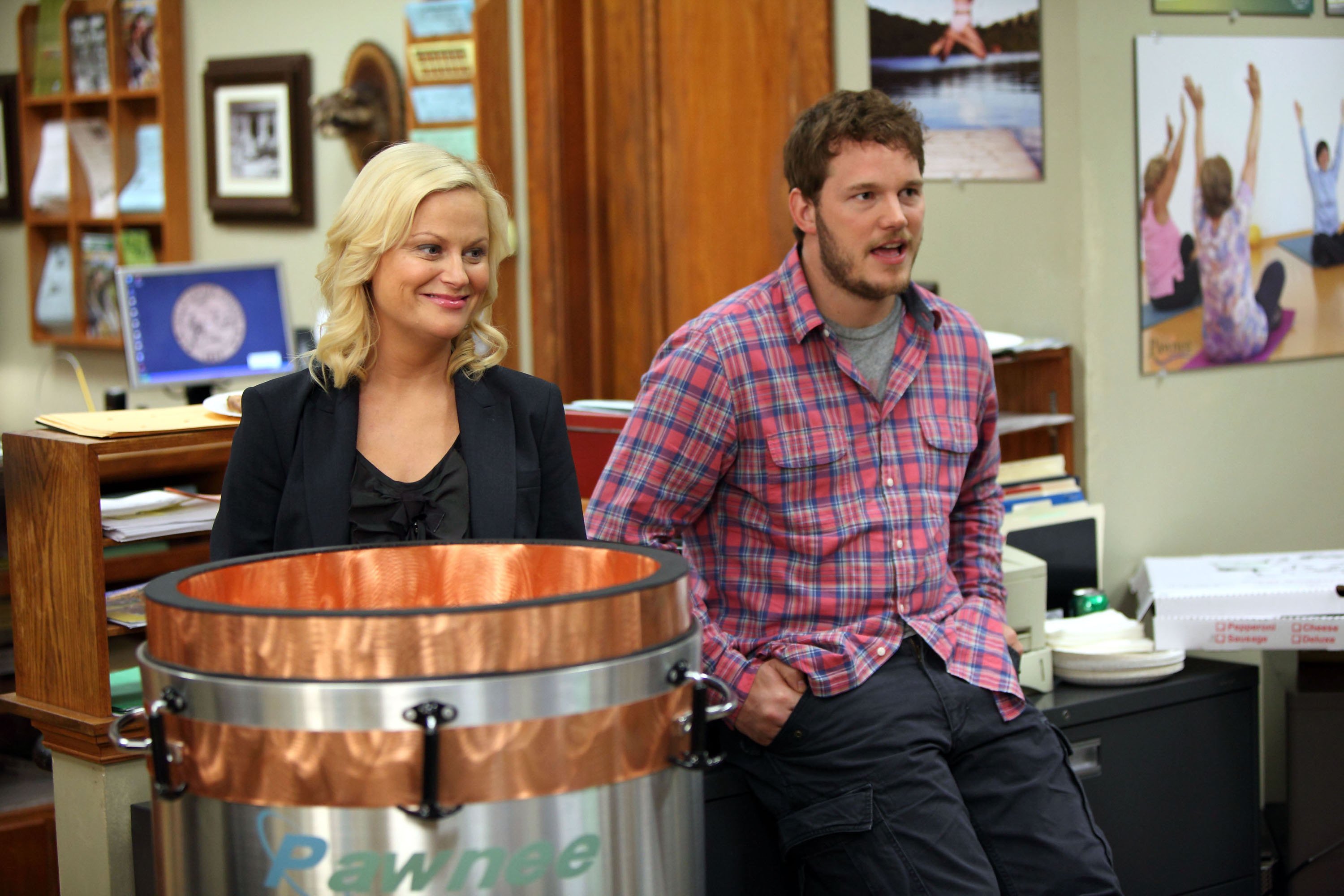 Chris Pratt sits on a desk next to Amy Poehler