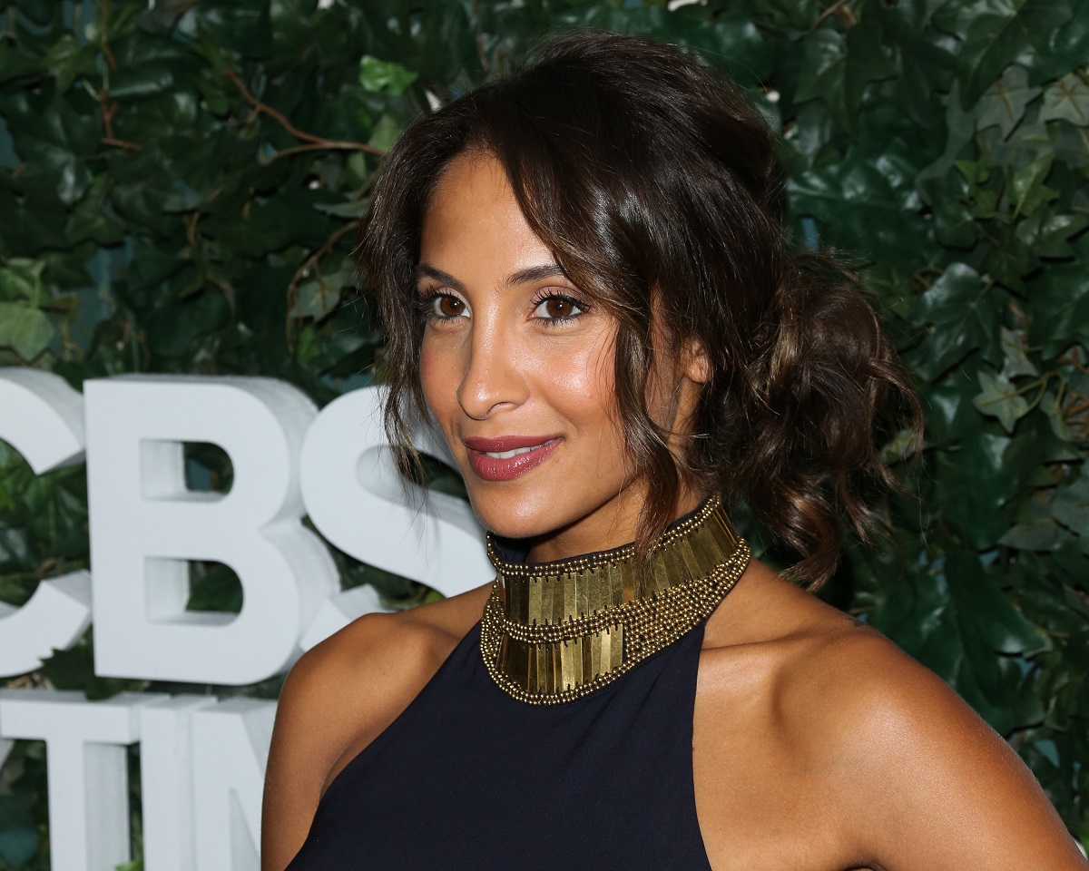 'The Young and the Restless' actor Christel Khalil wearing a blue and gold dress, stands in front of a hedge.