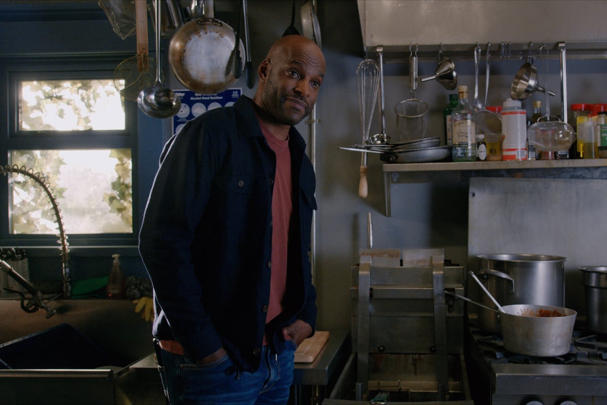 Colin Lawrence as Preacher leaning against a stove with his hands in his pocket in 'Virgin River'