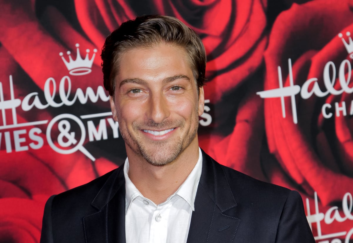 Daniel Lissing wearing a black suit and white shirt smiling in front of a red background.