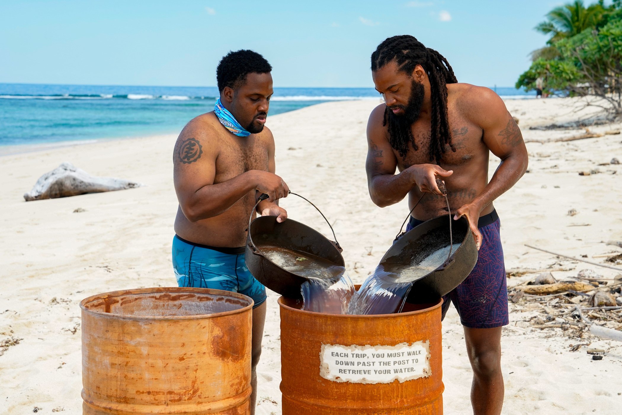 Deshawn Radden and Danny McCray, members of the 'Survivor' Season 41 cast, pouring water into a large barrel on the beach