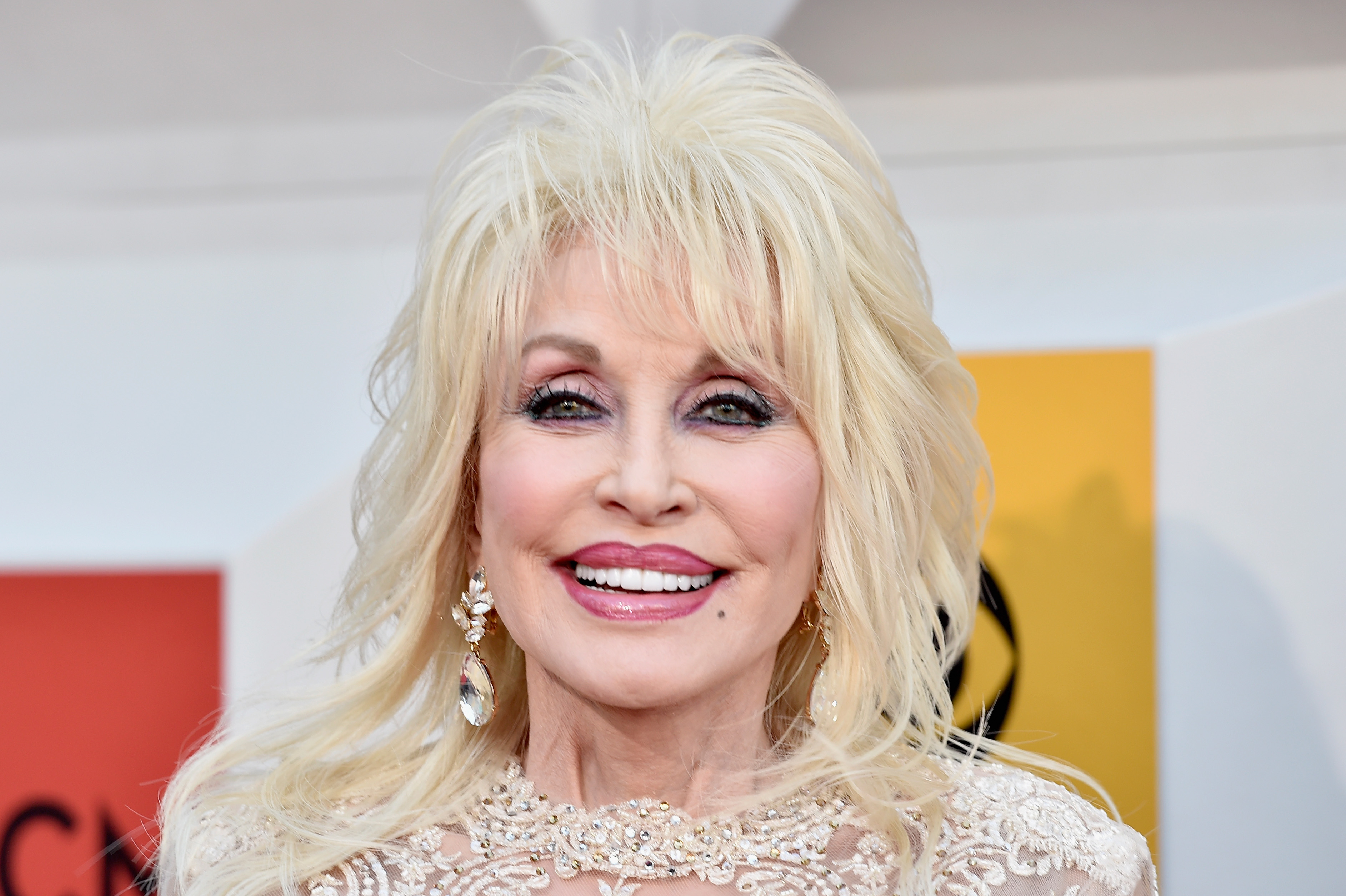 Dolly Parton wearing a white embroidered top and earrings in front of a red and yellow background.