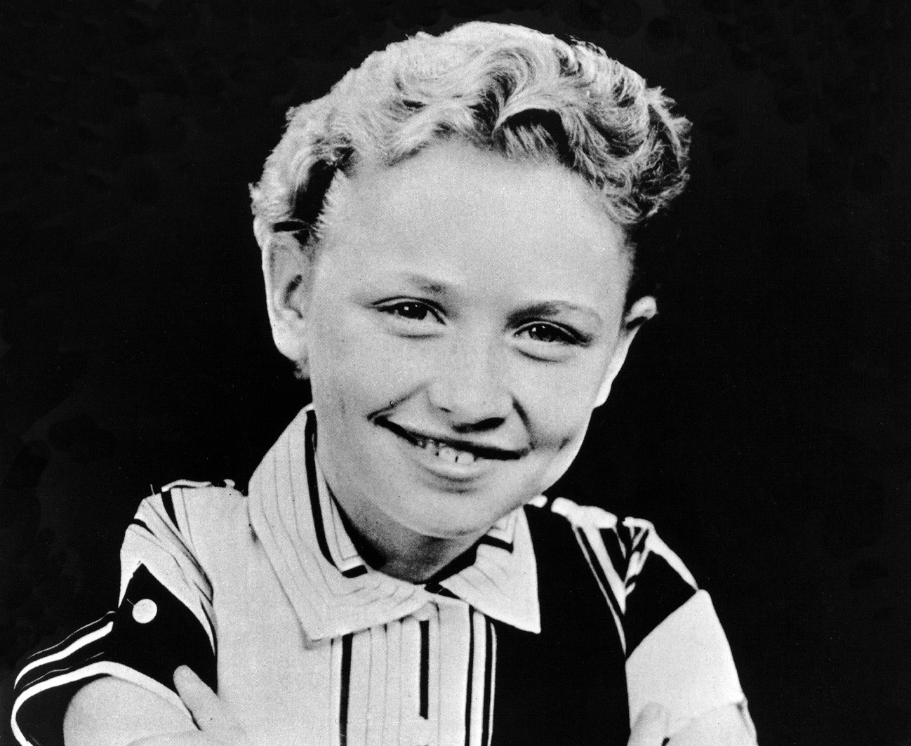 A black and white photo of Dolly Parton as a child sitting in front of a black background.