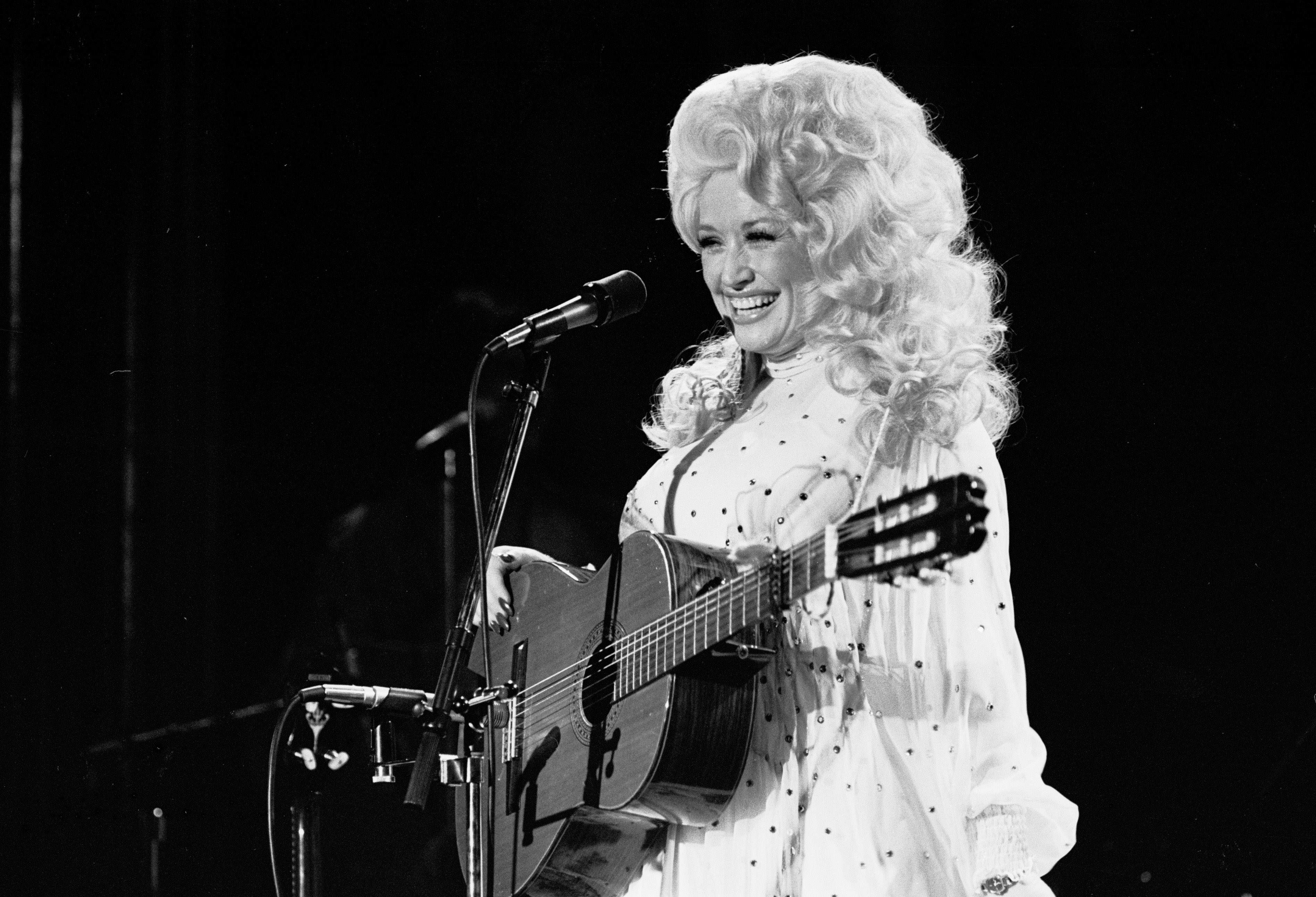 A black and white photo of Dolly Parton  in a white long sleeve dress. She holds her guitar and stands in front of a microphone.