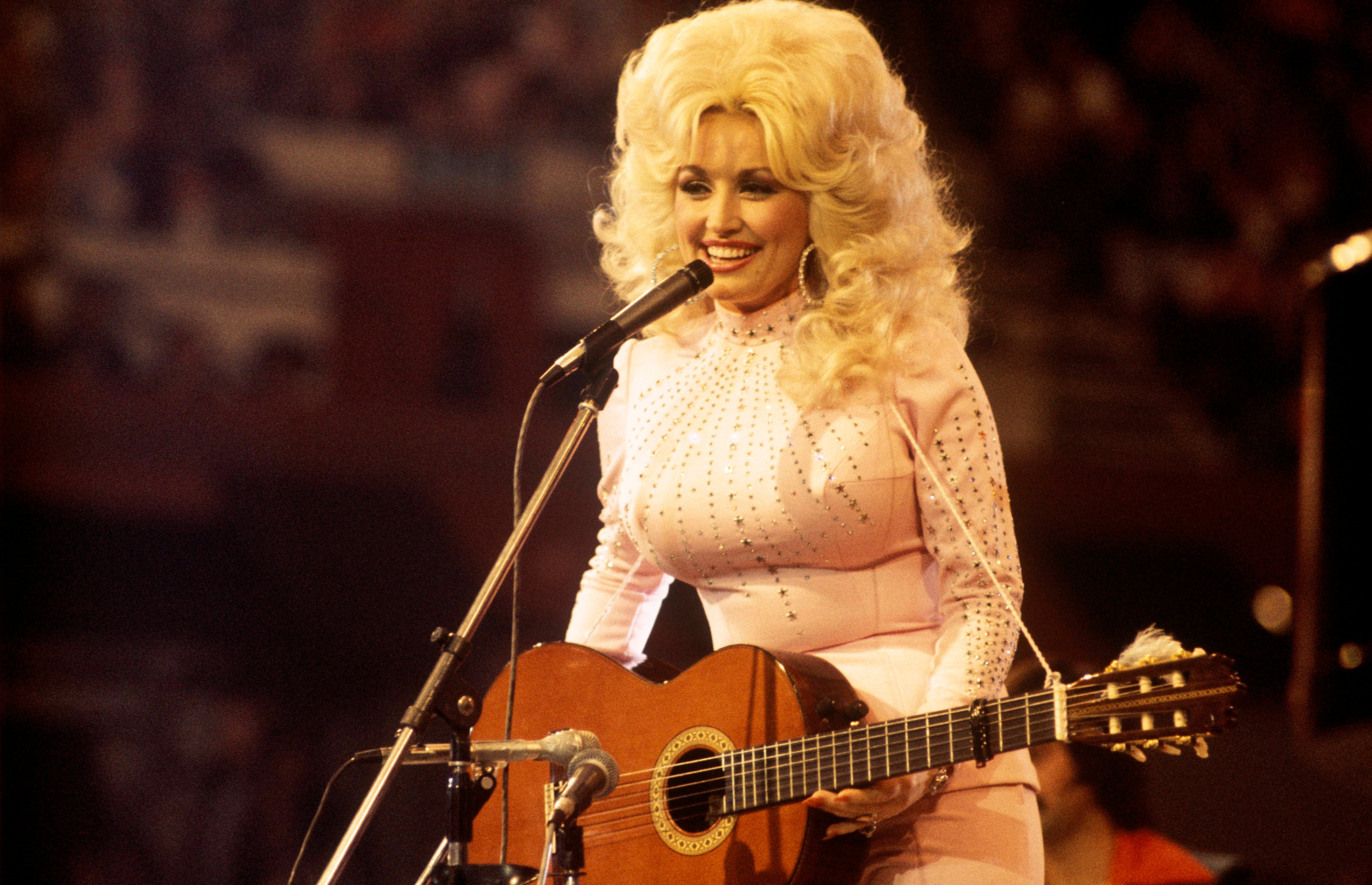 Dolly Parton wears a white sparkly shirt and holds a guitar. She stands in front of a microphone.