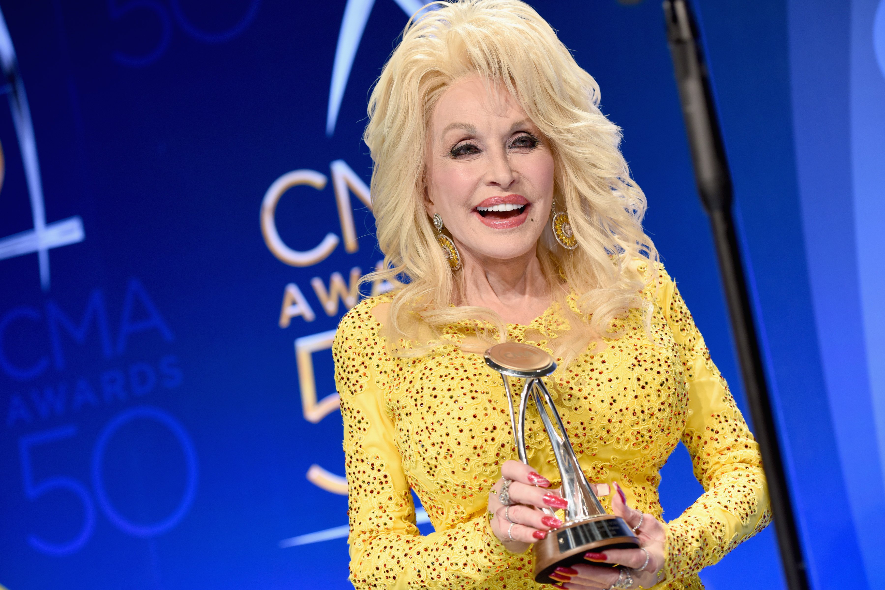 Dolly Parton wears a yellow jeweled dress while holding an award.
