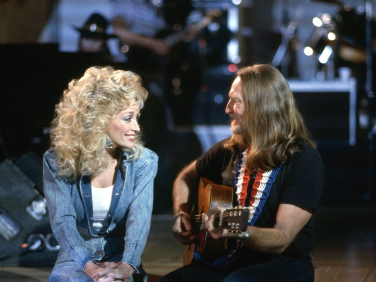 Dolly Parton (L) in denim, smiling at Willie Nelson (R) in a black tee and playing a guitar, also smiling at Parton