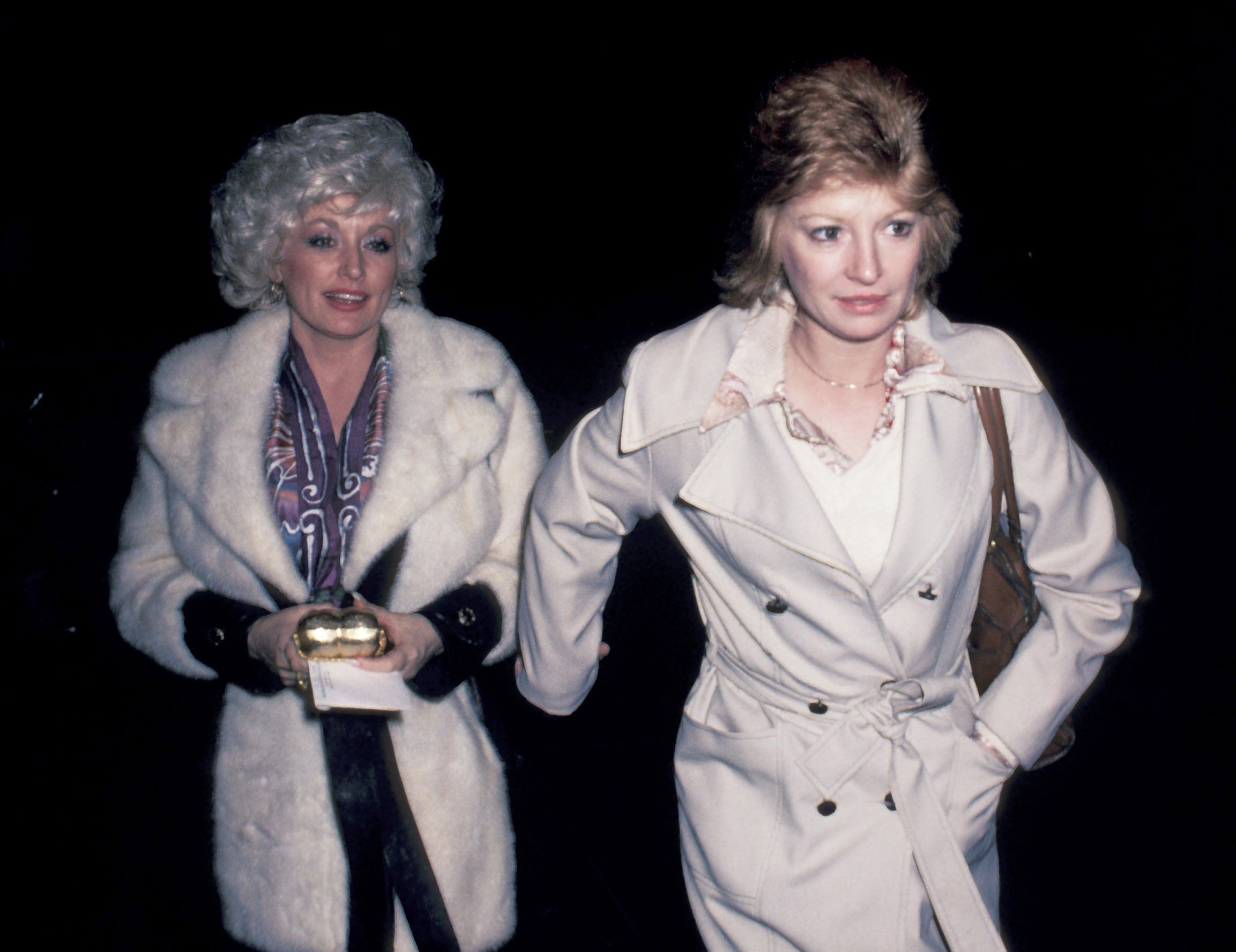Dolly Parton in a fuzzy white coat and Judy Ogle in a trench coat. They walk outside together at night.