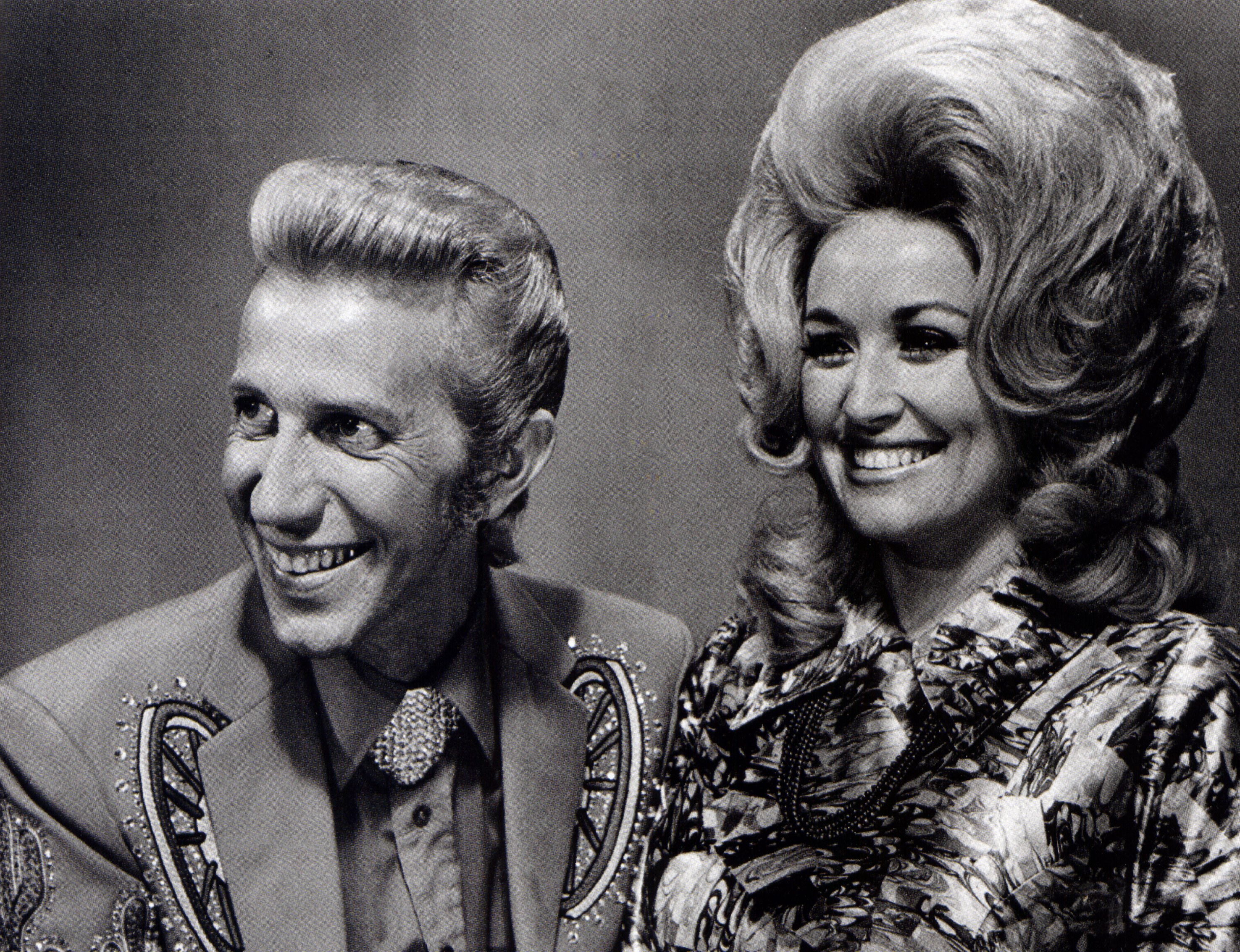 Dolly Parton and Porter Wagoner pose together, smiling, in a black and white photo.