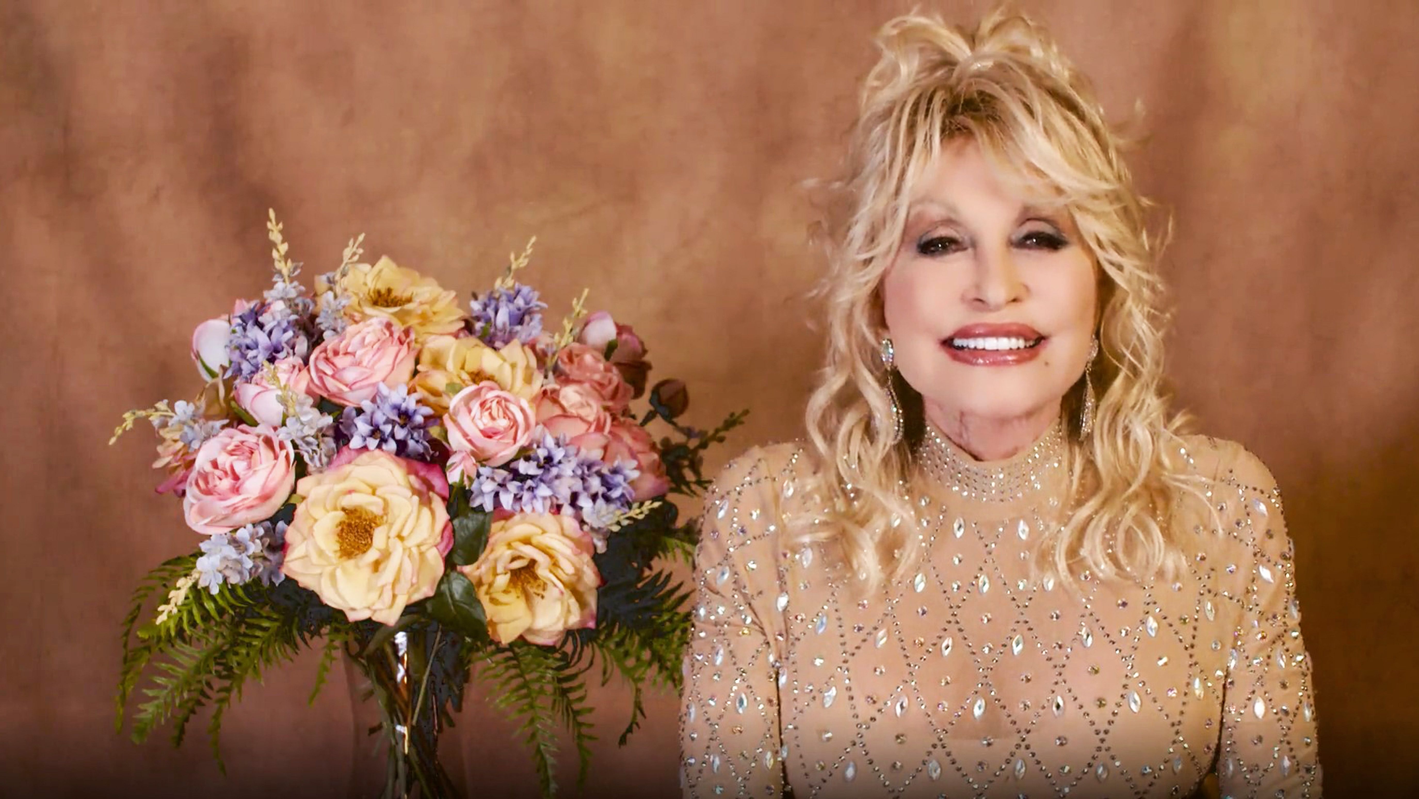 Dolly Parton smiles while wearing a jeweled dress and sitting in front of a bouquet of flowers. 