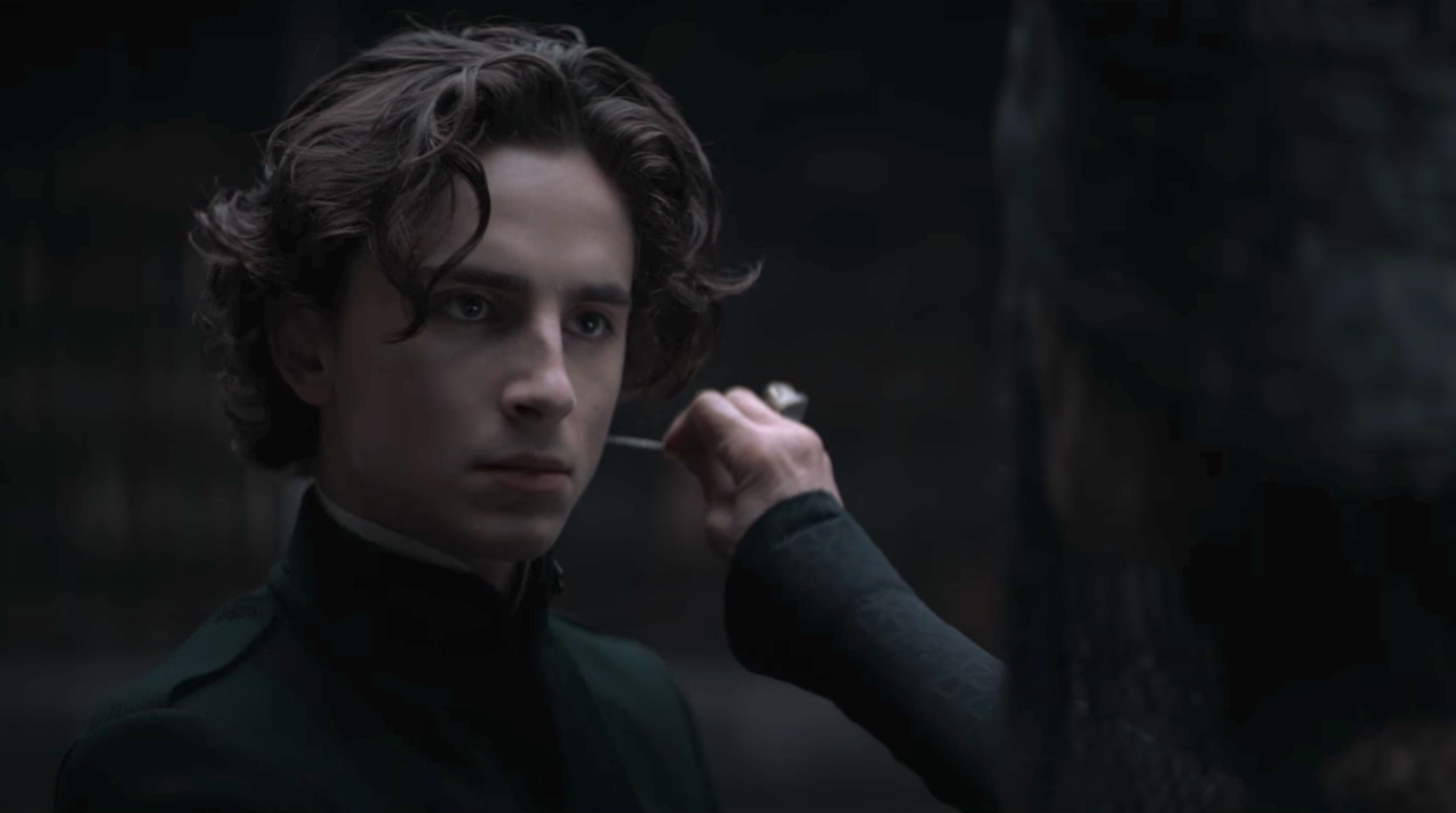 Timothée Chalamet and Charlotte Rampling in the 'Dune' Gom Jabbar scene. Chalamet looks at Rampling with a serious face as she holds a poisoned needle to his neck. He wears a green military-looking jacket with a high neck. Rampling wears all black and a black, beaded veil over her face. They're in a dimly lit library.