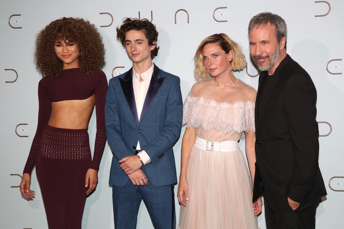 Denis Villeneuve and the main cast of Dune on the red carpet. L-R: Zendaya in a purple crop top and long skirt, Timothée Chalamet in a blue suit and white shirt, Rebecca Ferguson in a blush pink dress, and Denis Villeneuve in a black suit and shirt. They stand in front of a white backdrop that says 'DUNE' in gold lettering.