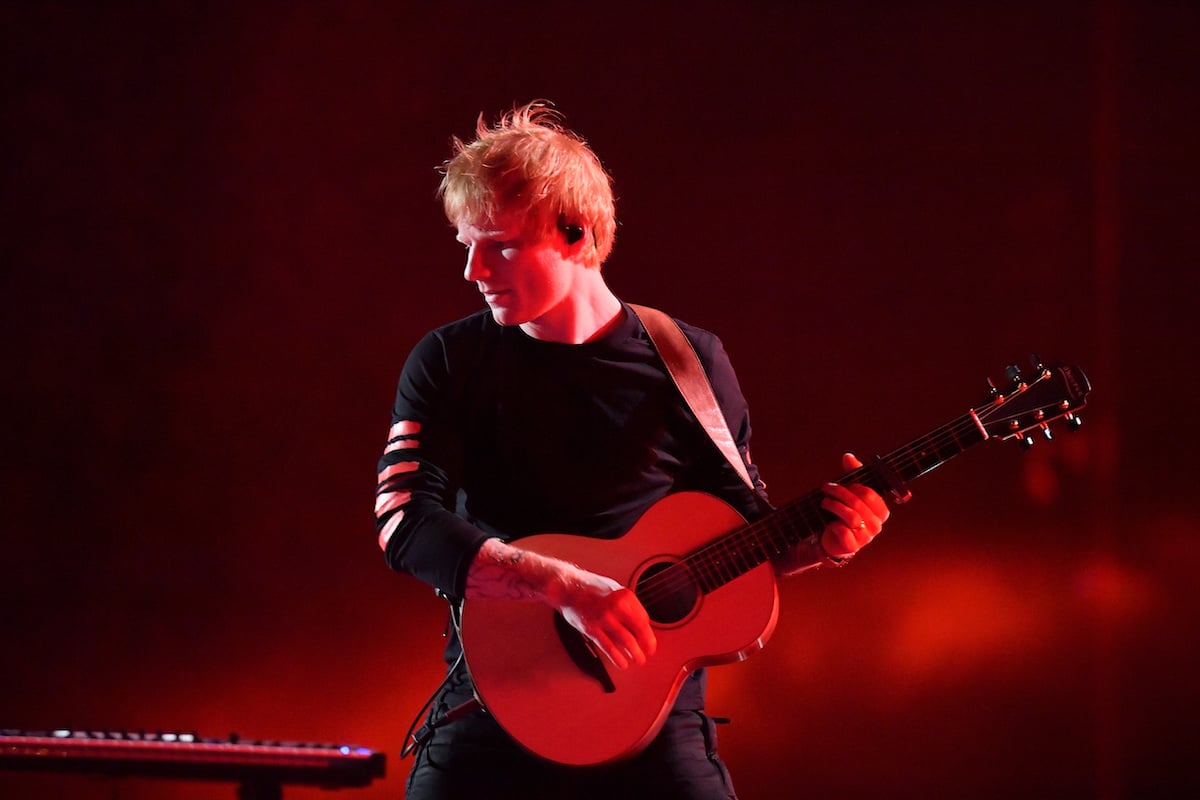 Ed Sheeran joue de la guitare sur scène sur fond rouge.