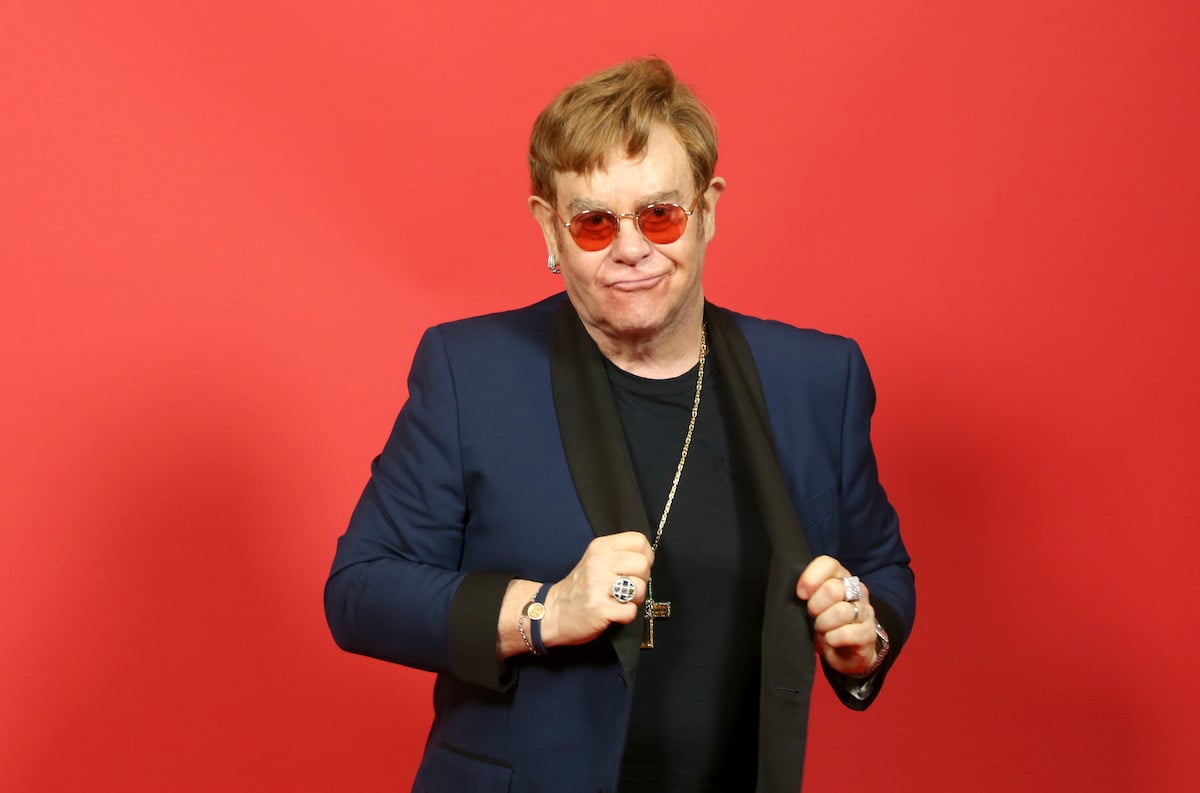 Elton John poses in front of a red background.