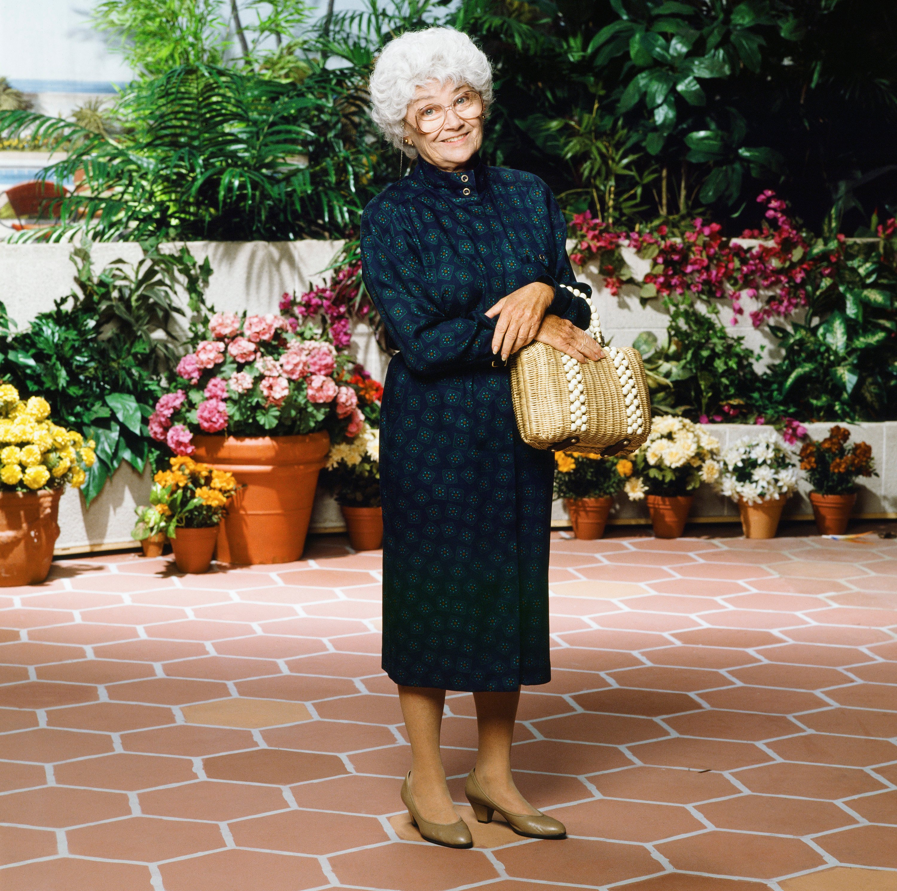 'The Golden Girls' actor Estelle Getty wearing a blue dress and glasses, and standing on a lanai.