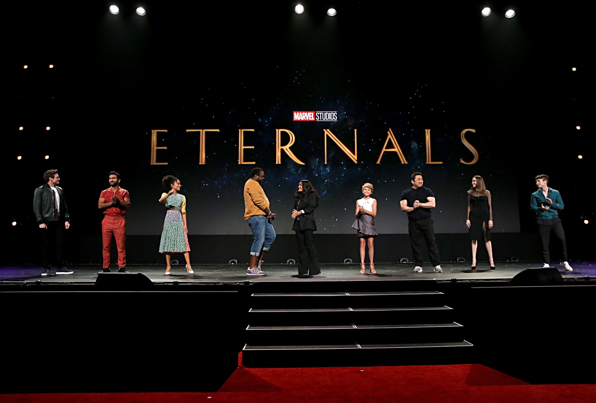 (L-R) Richard Madden, Kumail Nanjiani, Lauren Ridloff, Salma Hayek, Lia McHugh, Don Lee, Angelina Jolie, and Barry Keoghan of 'The Eternals,' which has a new trailer. They're standing on-stage and a logo for the film is behind them.