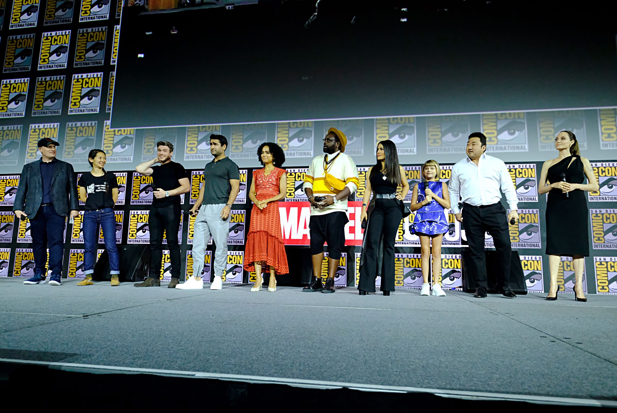 The cast and crew of Marvel's 'Eternals' stand onstage during a presentation at San Diego Comic-Con. They stand in front of a Marvel sign, a big screen, and posters for the convention. 'Eternals' will have two post-credits scenes.