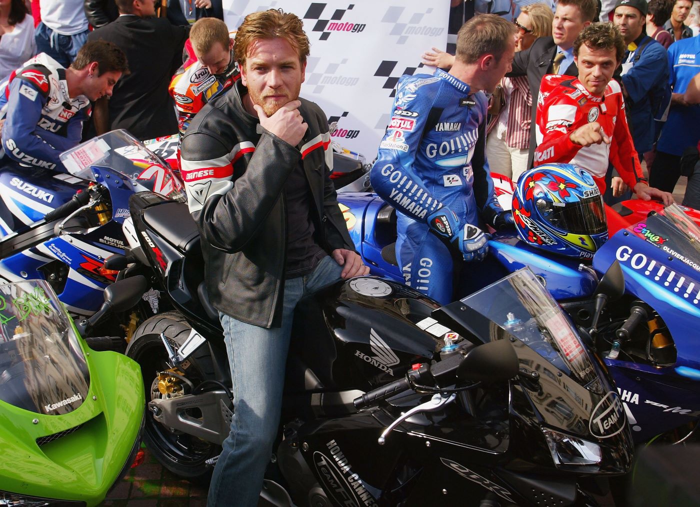 Ewan McGregor on a sports bike dressed in motorcycle gear surrounded by other motorcycles and their riders.