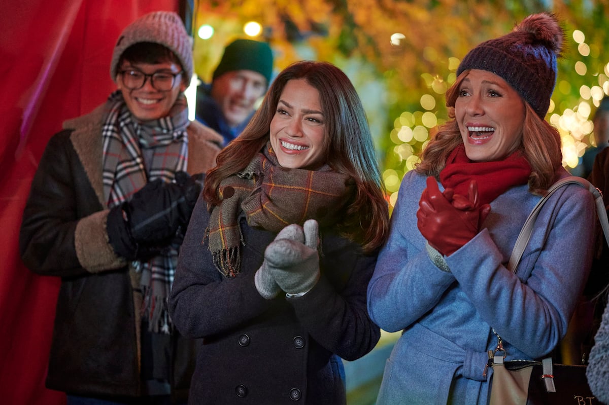 Three people dressed in winter clothes smiling and clapping in the Hallmark Christmas movie 'Five Star Christmas'