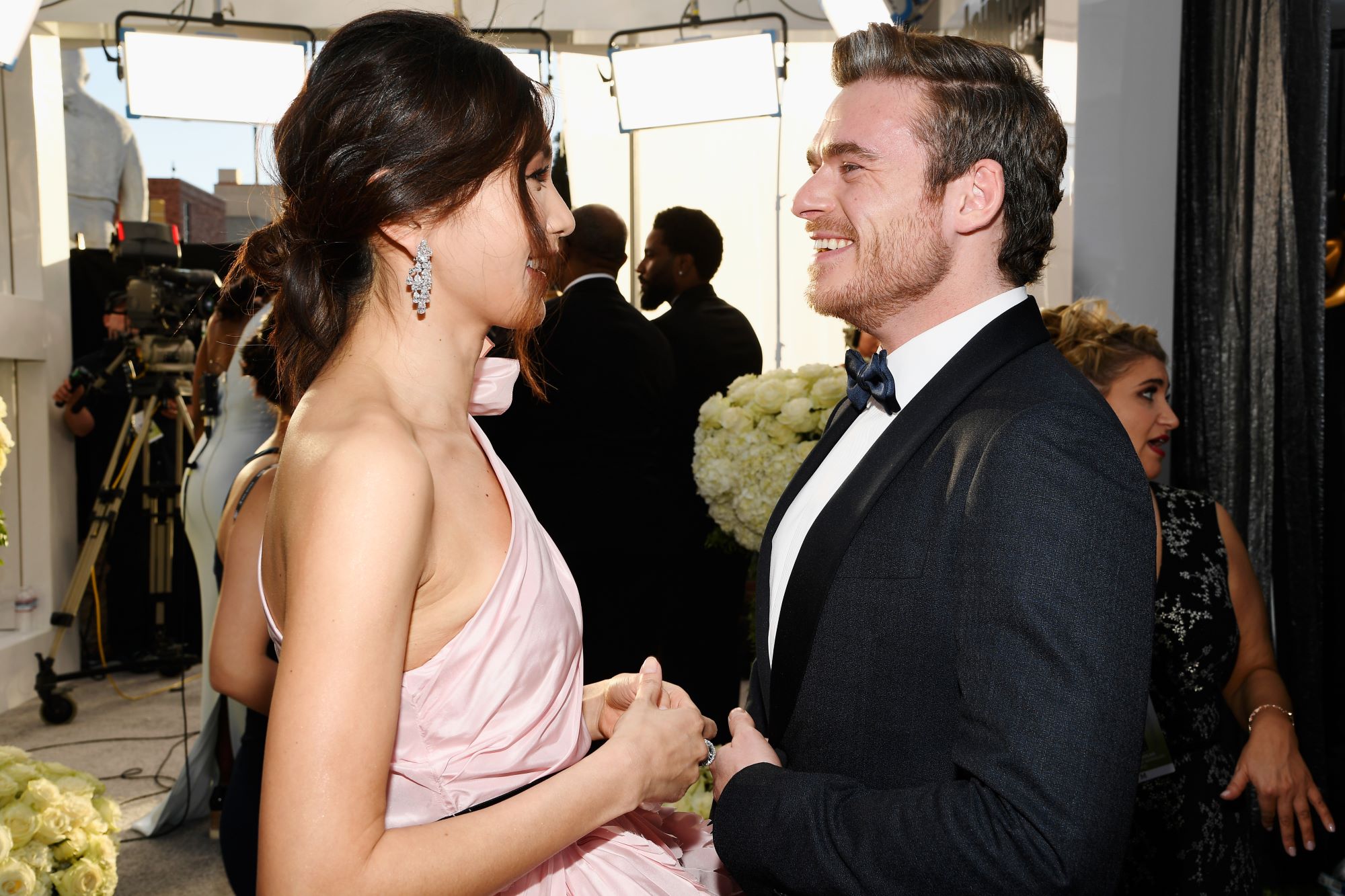 Marvel's 'Eternals' stars Gemma Chan and Richard Madden talk on the red carpet. Chan wears a light pink, one-shoulder dress and dangly earrings. Madden wears a black suit over a white button-up shirt and a black bow tie.