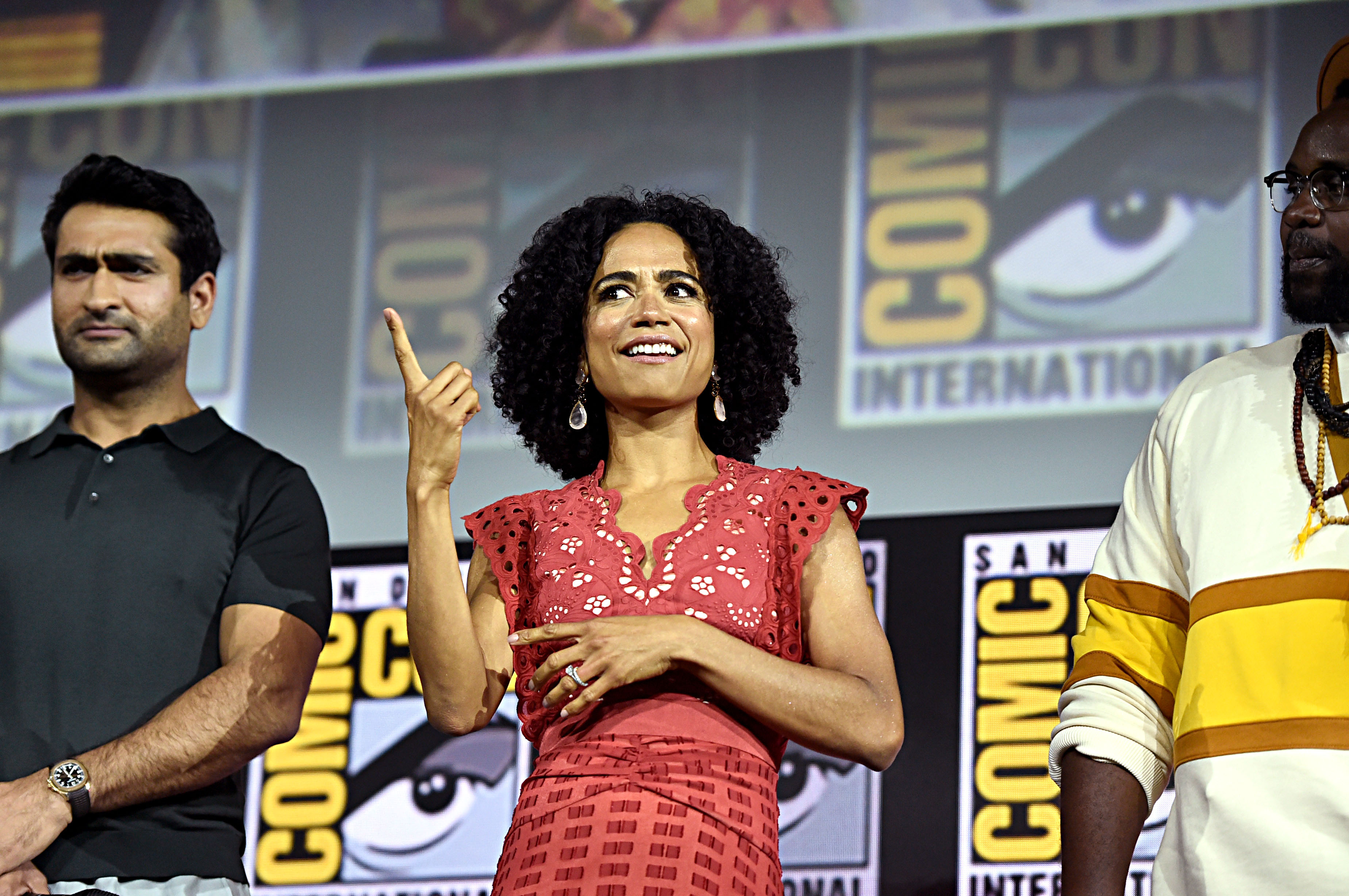 'Eternals' stars Kumail Nanjiani, Lauren Ridloff, and Brian Tyree Henry gather onstage. Nanjiani wears a black polo shirt. Ridloff wears a red dress. Henry wears a white shirt with a yellow stripe.