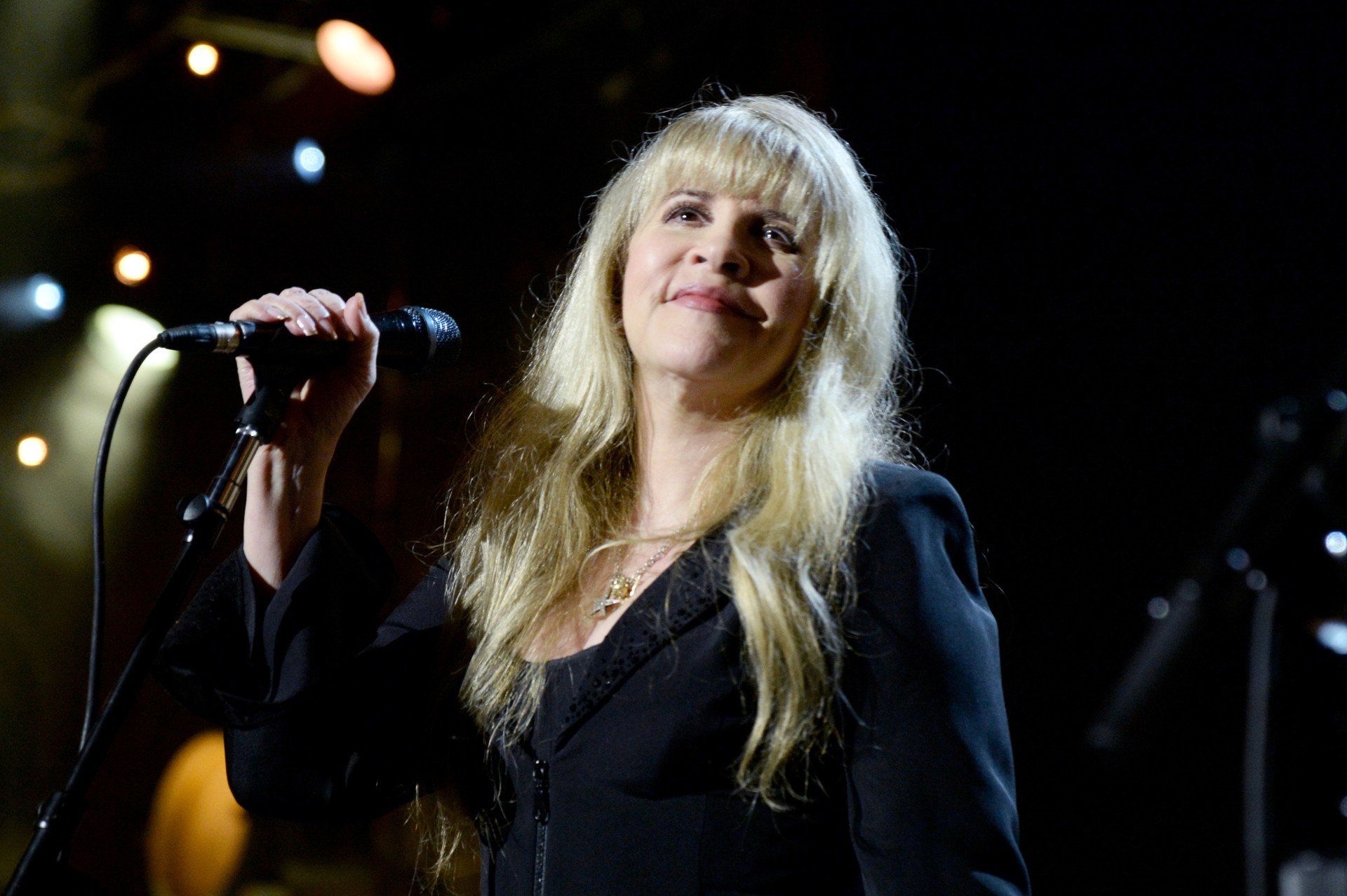 Stevie Nicks stands in front of a microphone while wearing a black outfit with flared sleeves