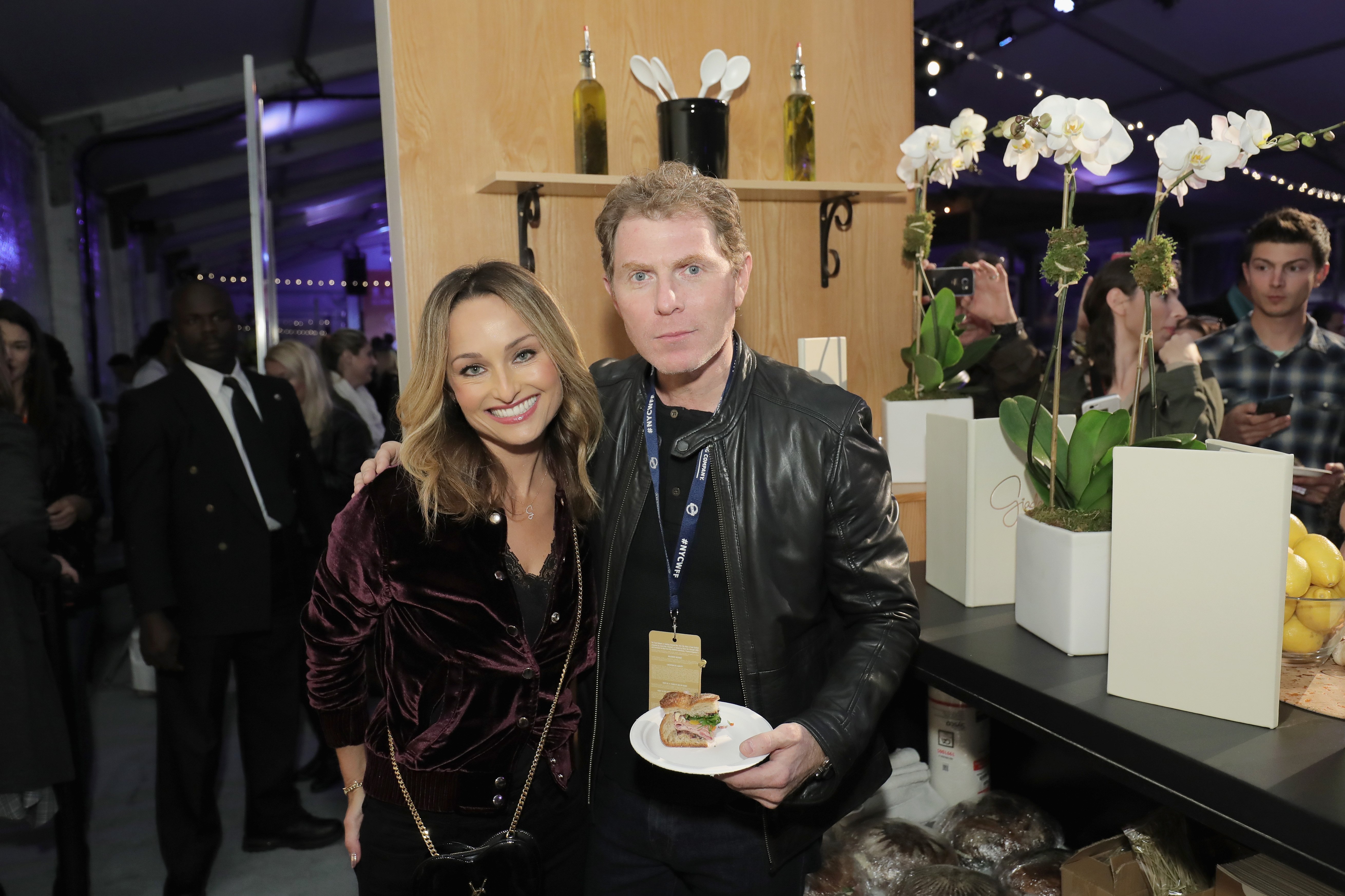 Bobby Flay and Giada De Laurentiis pose at an event while Bobby Flay holds a plate of food.