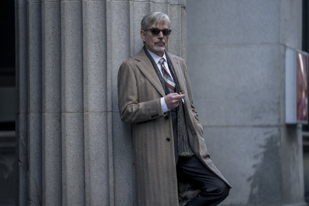 Billy McBride wears a suit, coat and sunglasses in Goliath Season 4. He is leaning against a wall smoking a cigarette.