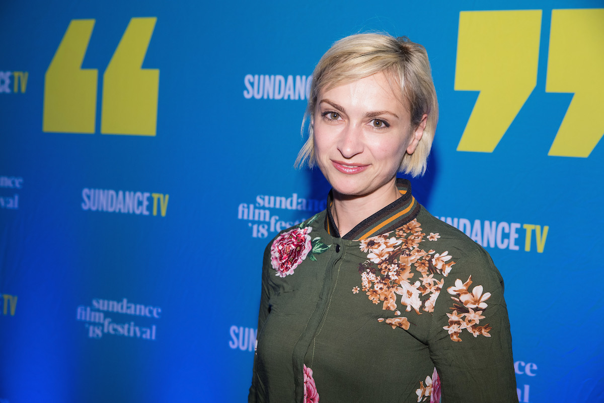 Halyna Hutchins on a red carpet at Sundance. She wears a green floral dress and stands in front of a blue backdrop with white and yellow lettering.