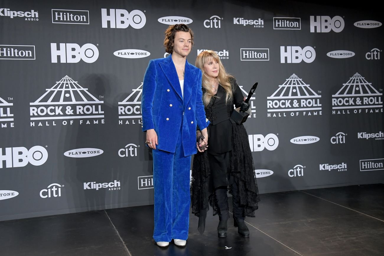 Harry Styles in a blue suit holds hands with Stevie Nicks in a black dress.