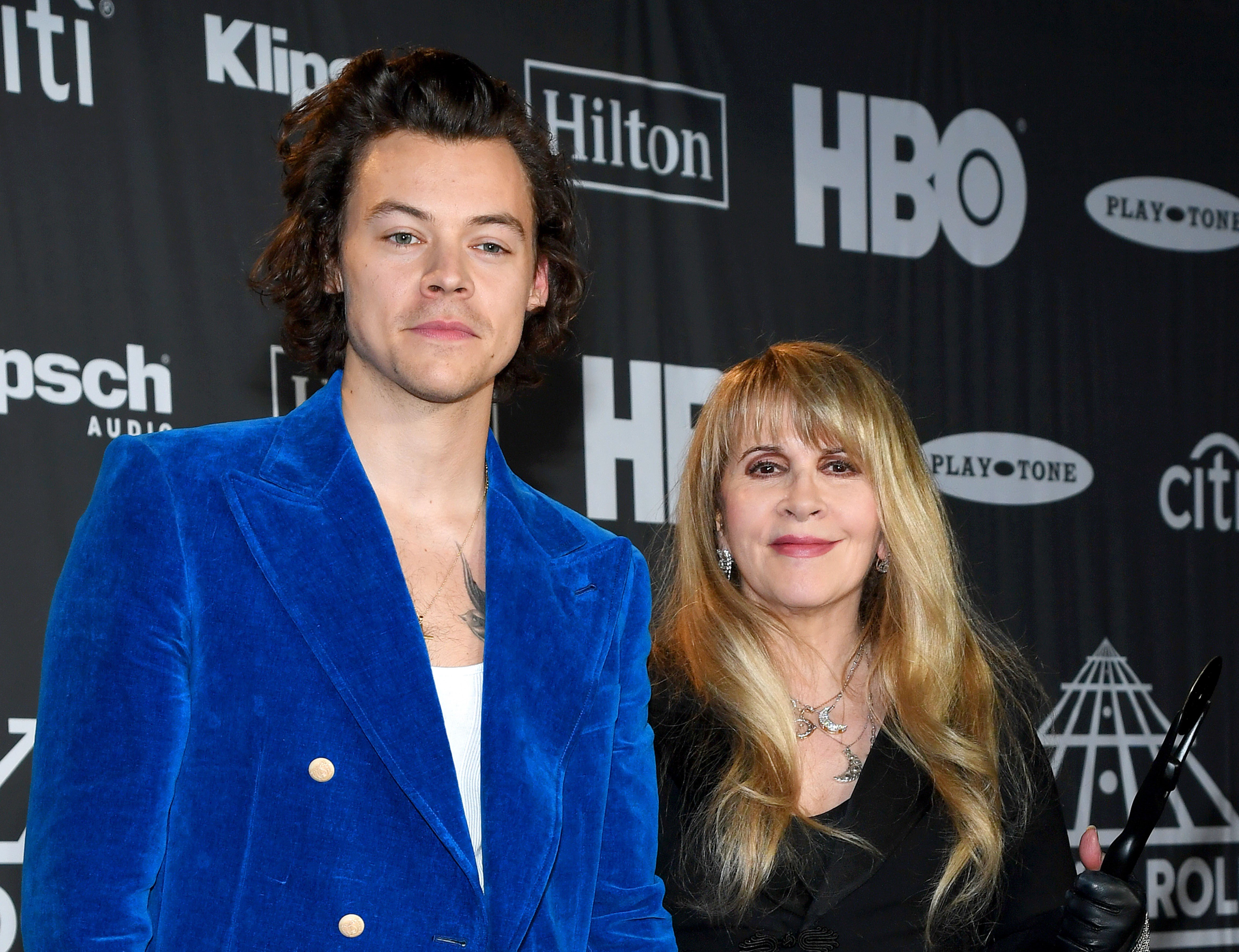 Harry Styles wears a blue suit and Stevie Nicks wears a black dress. They stand in front of a black background.
