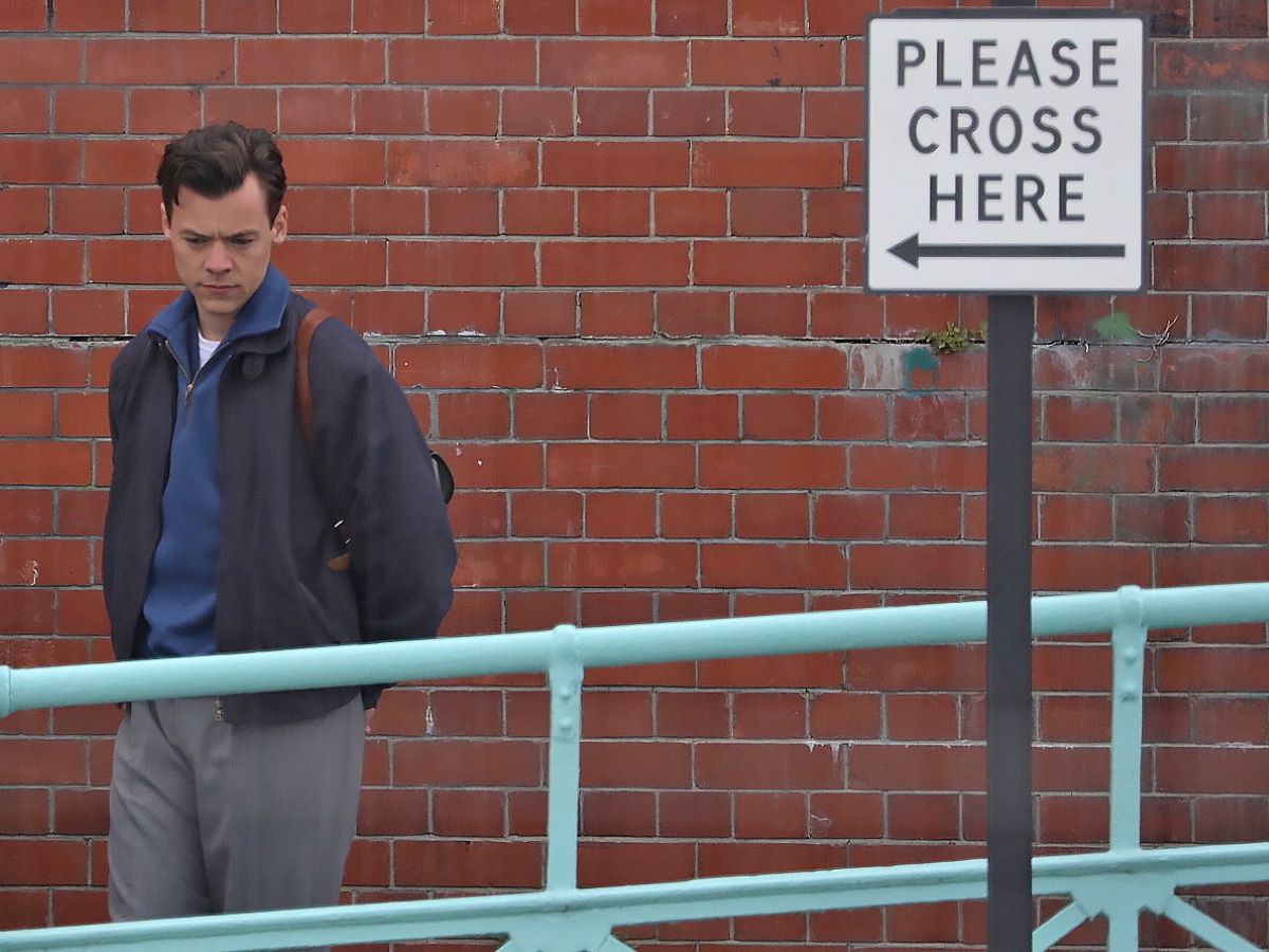 Clean cut Harry Styles on the set of a movie, standing next to a 'please cross here' sign