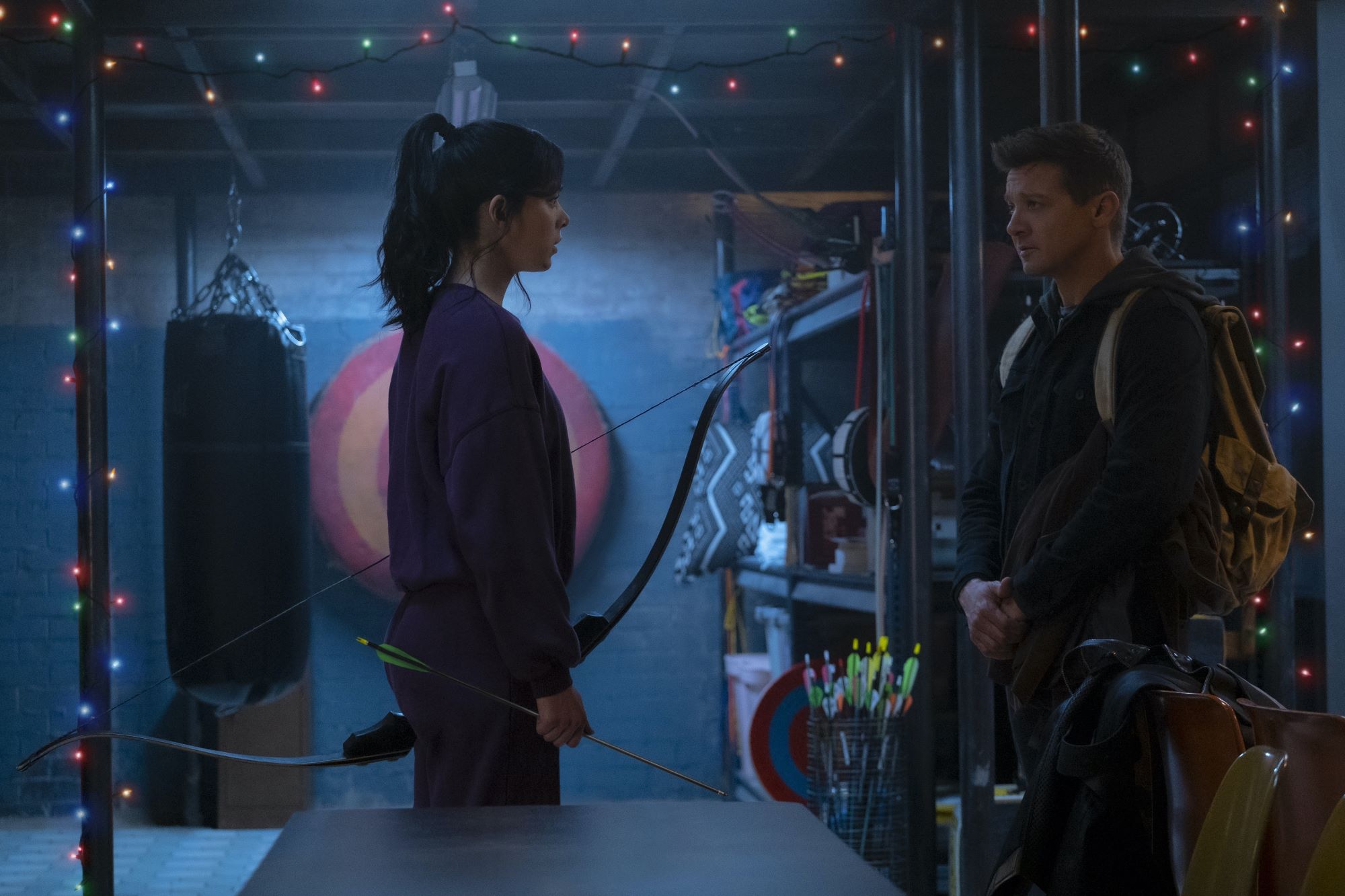 'Hawkeye' stars Hailee Steinfeld and Jeremy Renner, in character as Kate Bishop and Clint Barton, stand in a training room with Christmas lights. Steinfeld wears a purple sweater and purple sweatpants and holds a bow and arrow. Renner wears a dark jacket and a backpack.