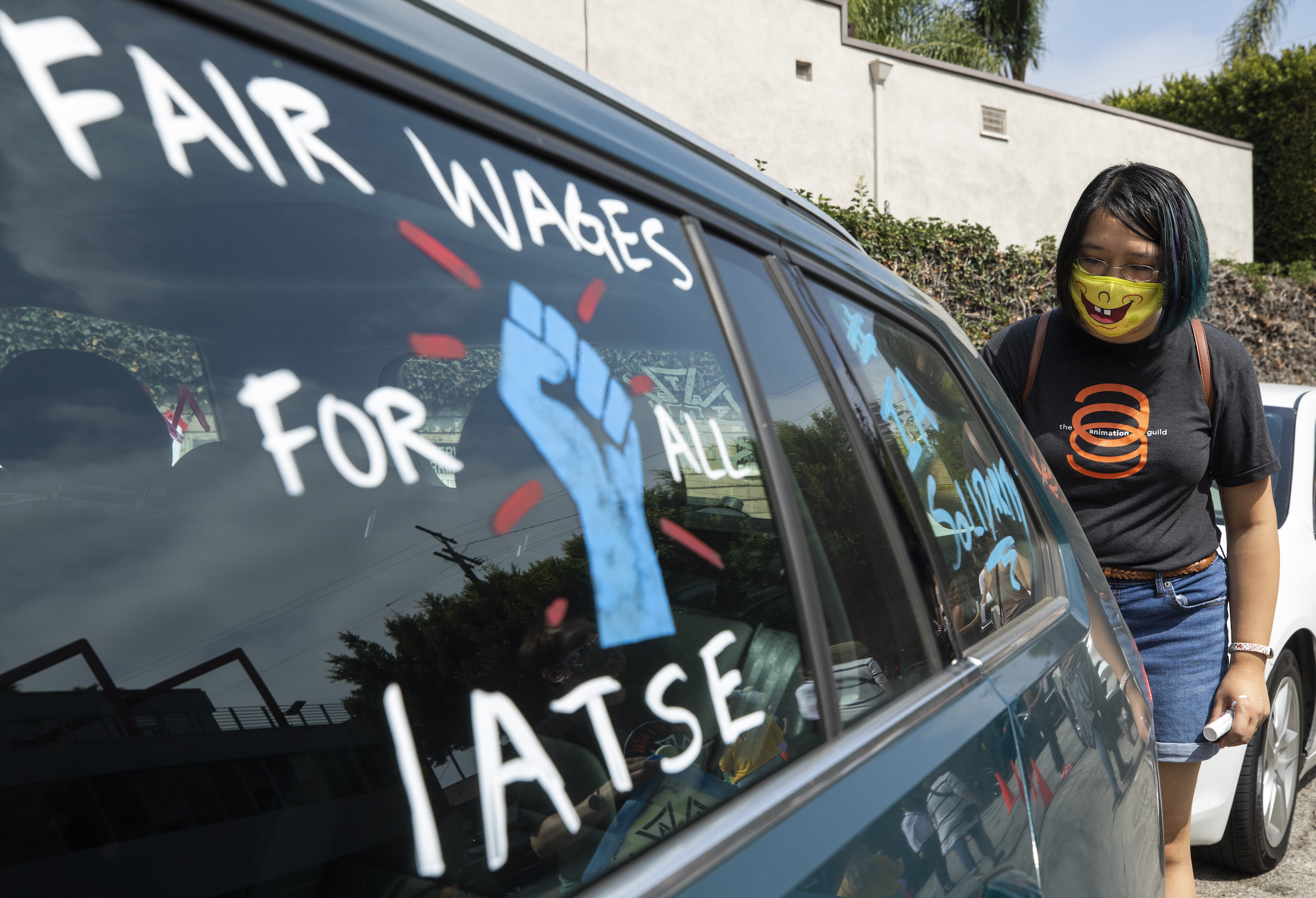 IATSE strike rally in Los Angeles. A Hollywood crew member stands by a car that says 'FAIR WAGES FOR ALL IATSE' and '#IASOLIDARITY' in the windows.