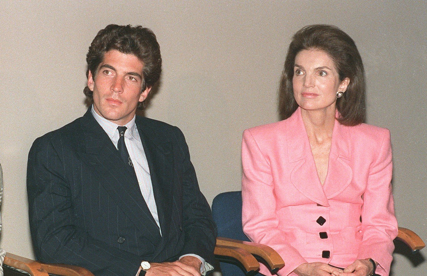 JFK Jr. wears a black suit and Jackie Kennedy Onassis wears a pink blazer. They sit next to each other. 