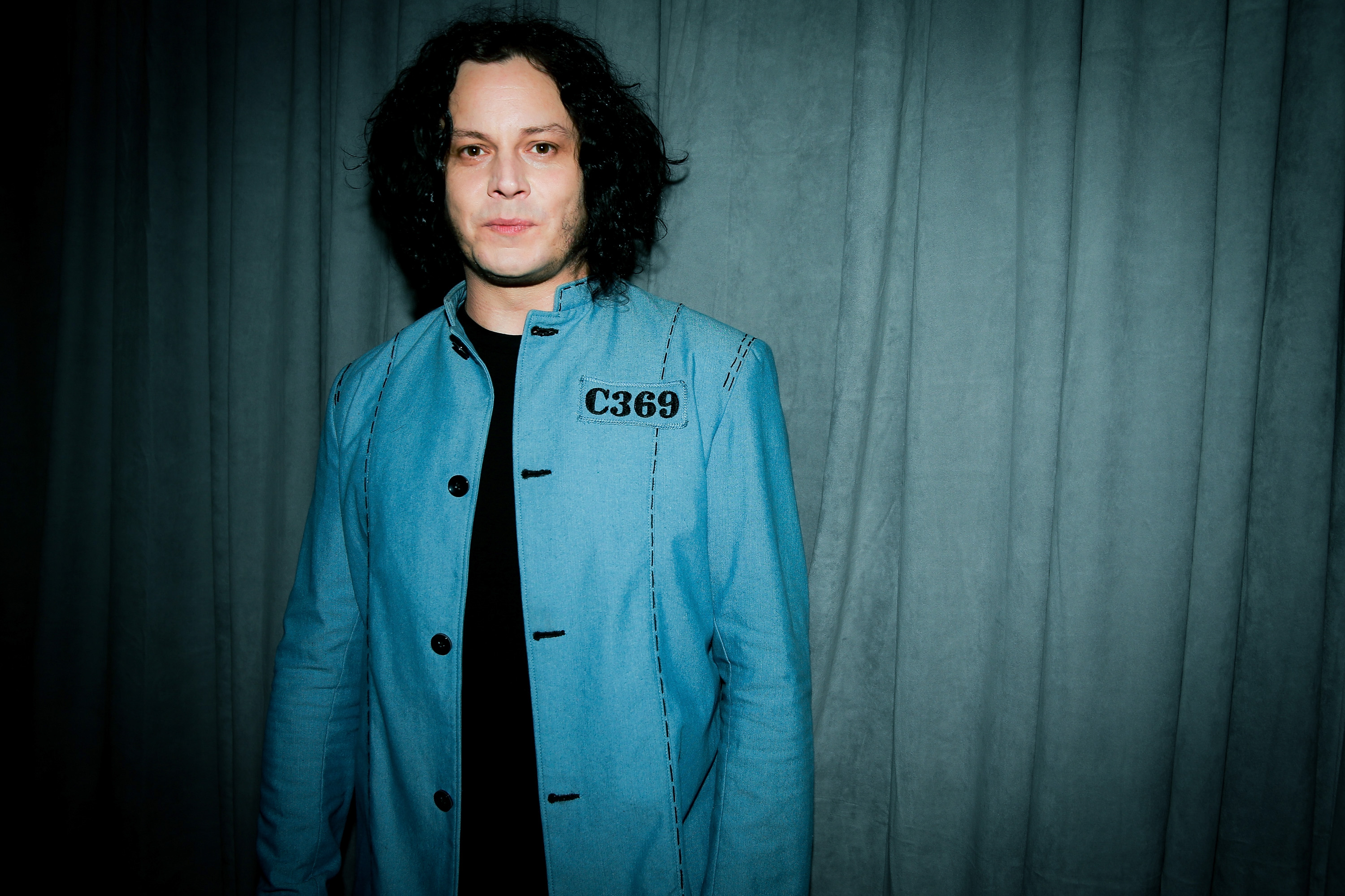 Jack White wears a black shirt and light blue jacket against a blue curtain background. 