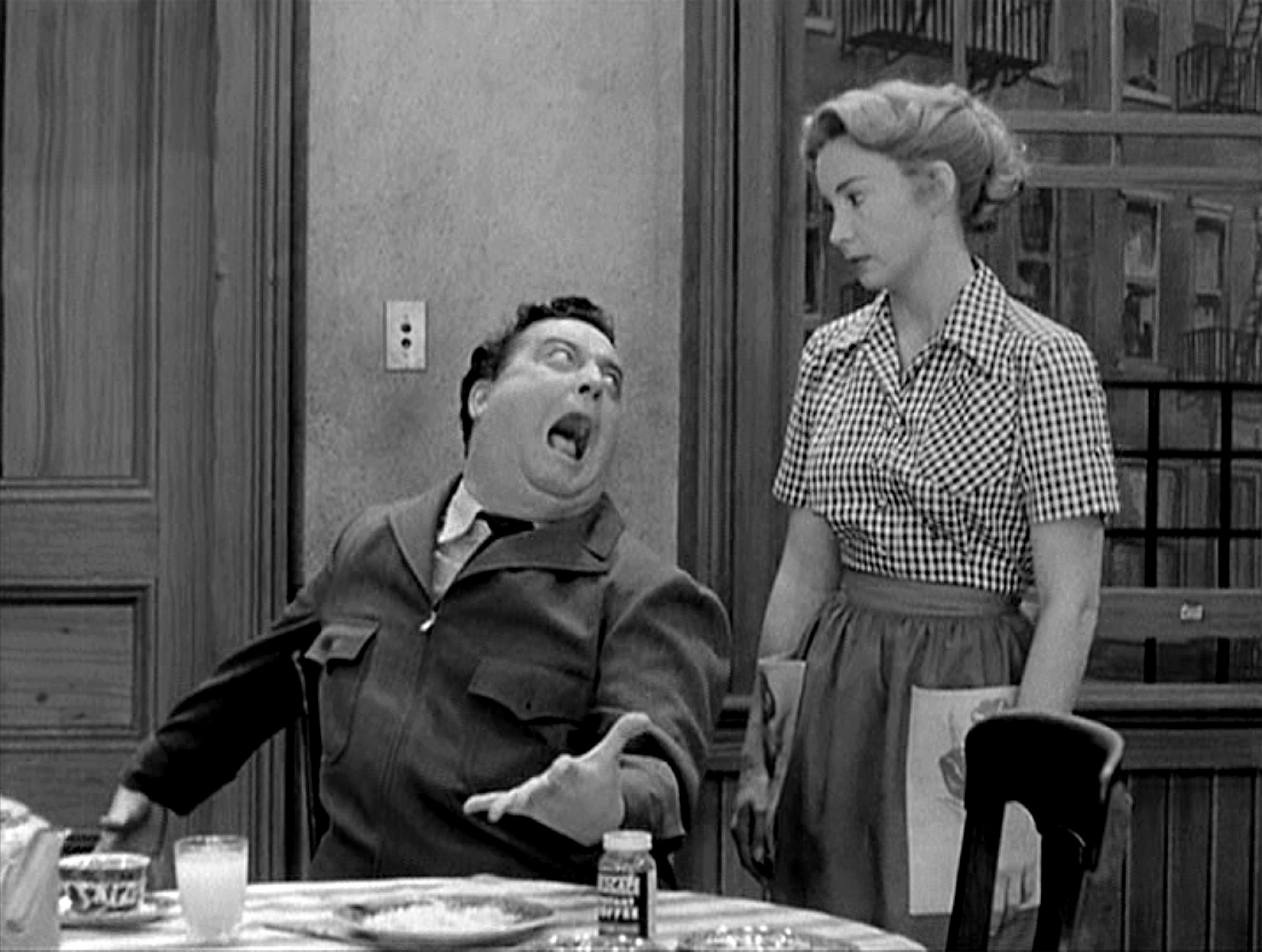 Jackie Gleason sits at a table and Audrey Meadows stands next to him on 'The Honeymooners' set.