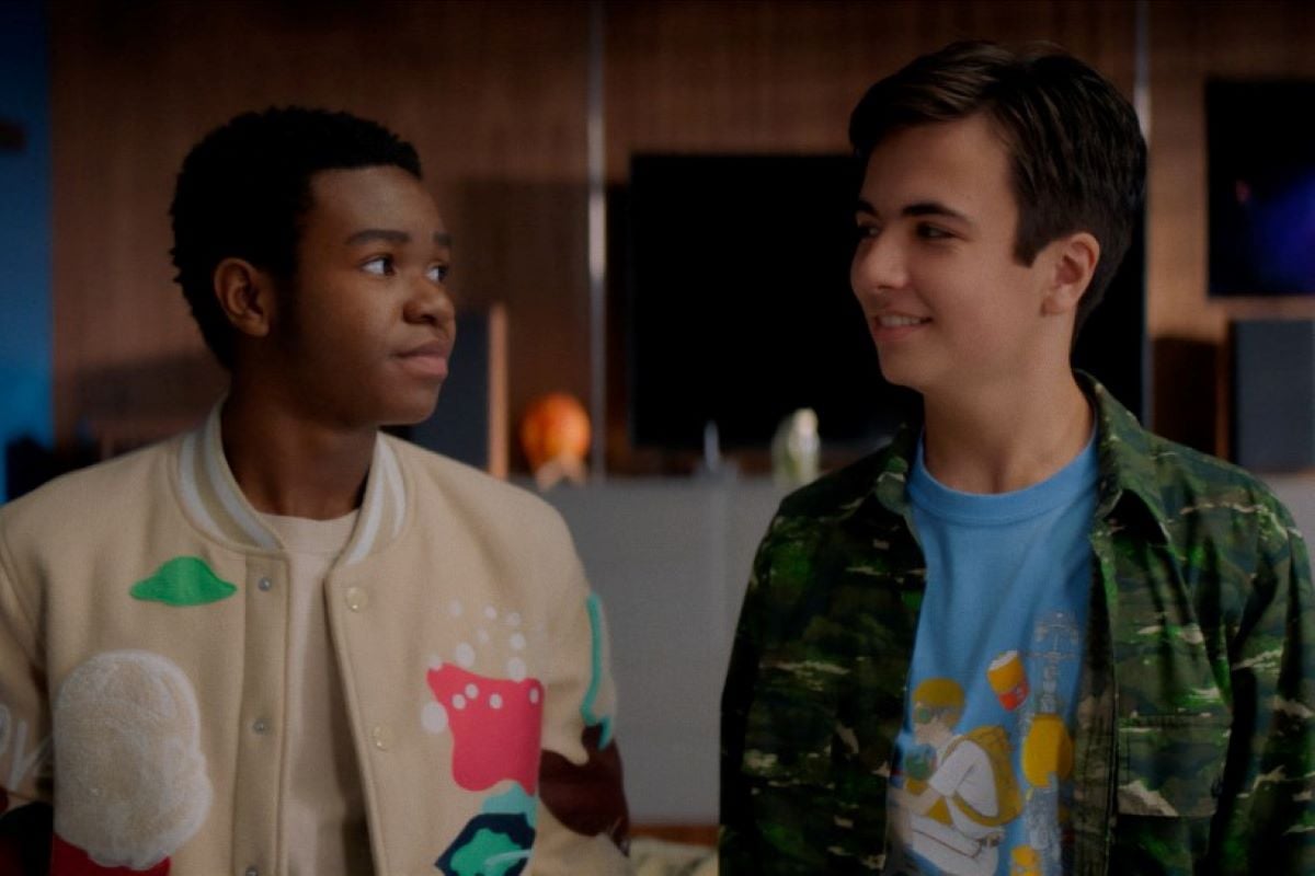 'Stargirl' Season 2 actors Alkoya Brunson and Trae Romano, in character as Jakeem Thunder and Mike Dugan, stand side by side and look at each other. Brunson wears a tan jacket with colorful patches on it. Romano wears a camo long-sleeved shirt over a blue shirt. Jakeem in the wielder of Thunderbolt.